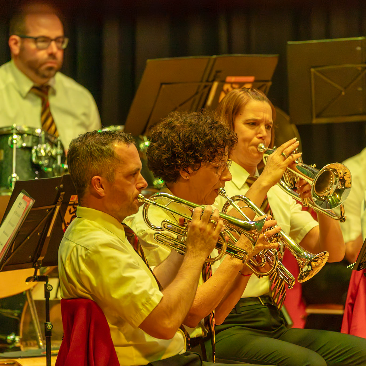 Eine Blaskapelle spielt auf der Bühne.  Frau [Name der Frau], die Trompete spielt, und Herr [Name des Mannes], der ein Flügelhorn spielt, sind konzentriert auf ihre Instrumente.  Die Musikerinnen und Musiker tragen einheitliche, helle Hemden und dunkle Hosen.  Die Musik erzeugt eine lebendige Atmosphäre.  Der Fokus liegt auf der musikalischen Leistung der Kapelle.