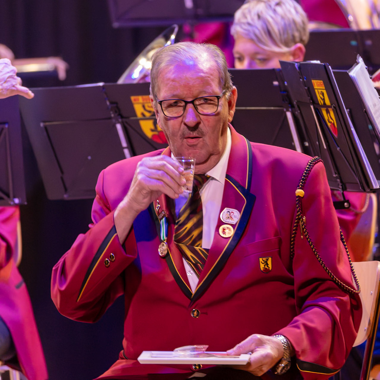 Herr [Name des Mannes], Dirigent/Mitglied der Blaskapelle, genießt einen kleinen Schluck während einer Pause der Blasmusik-Vorstellung.  Er hält ein kleines Glas in der Hand und scheint entspannt und zufrieden zu sein.  Die anderen Musiker der Kapelle sind im Hintergrund zu sehen, konzentriert auf ihre Instrumente.  Die Atmosphäre ist professionell und feierlich.