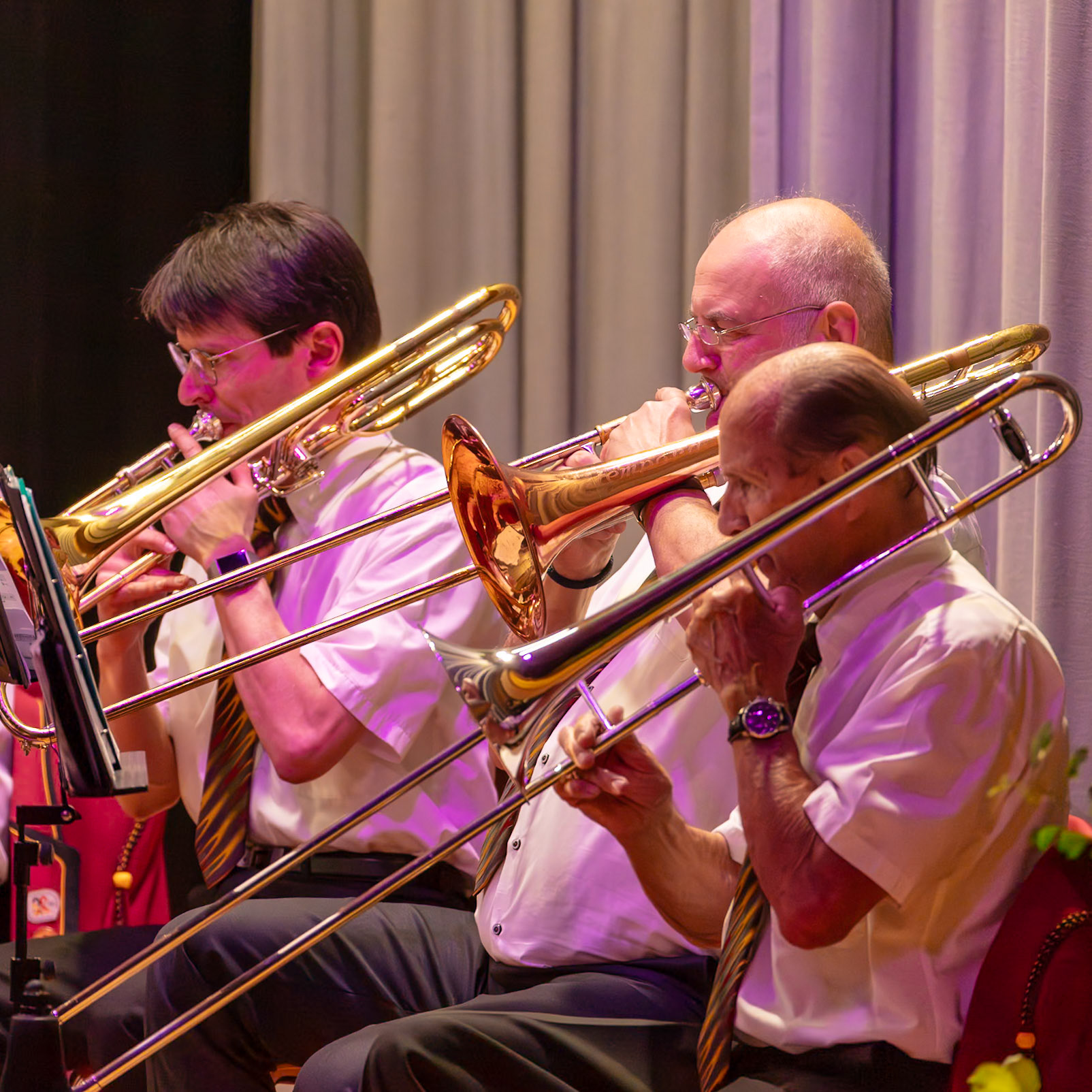 Auf der Bühne spielen drei Musiker Trompeten.  Der Mann in der Mitte, mit Brille, spielt eine Posaune.  Die Musiker konzentrieren sich auf die Ausführung ihrer Musikstücke.  Es ist ein Konzert oder eine ähnliche Veranstaltung, bei der ein Blasorchester spielt.