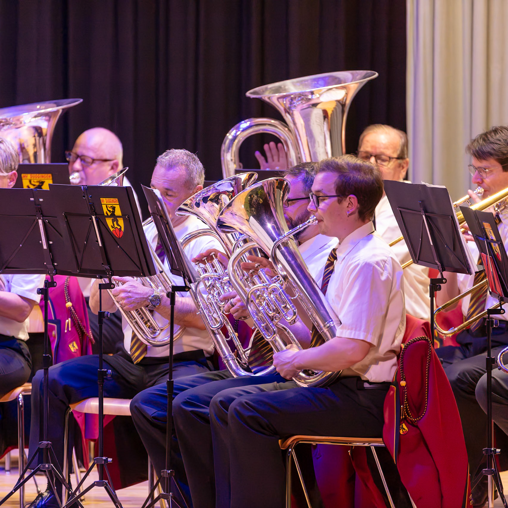 Ein Blasorchester spielt auf der Bühne.  Die Musiker, vermutlich Mitglieder des Blasorchesters "MV Duggen",  spielen auf ihren jeweiligen Instrumenten,  Trompeten und Tuba/Euphonien.  Sie tragen einheitliche weiße Hemden und dunkelblaue Hosen.  Die Atmosphäre ist konzentriert und professionell, während die Musiker ihre Musik darbieten.