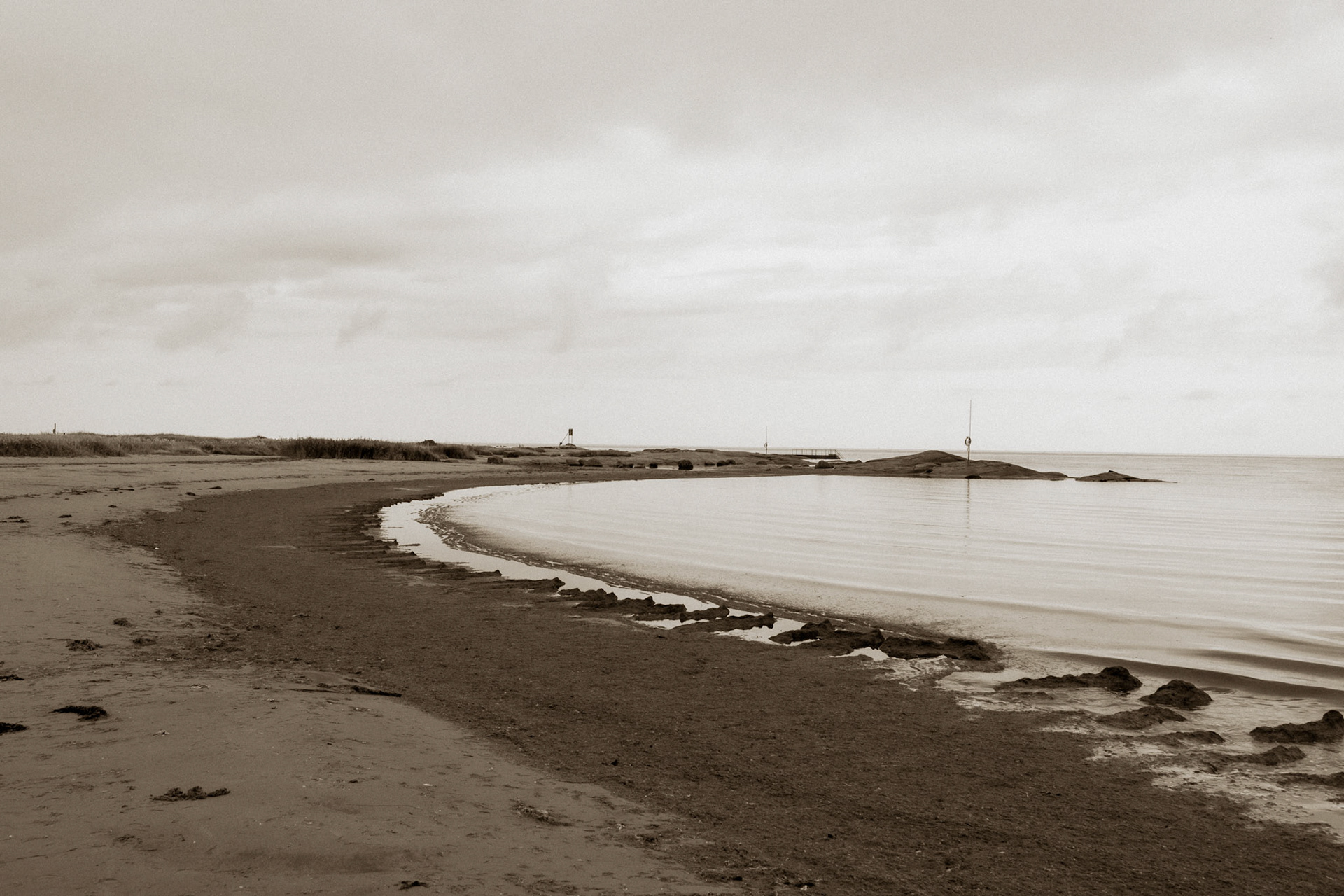 A serene coastal scene under a cloudy sky, featuring a calm sea meeting a sandy shore with gentle waves lapping at the edge.