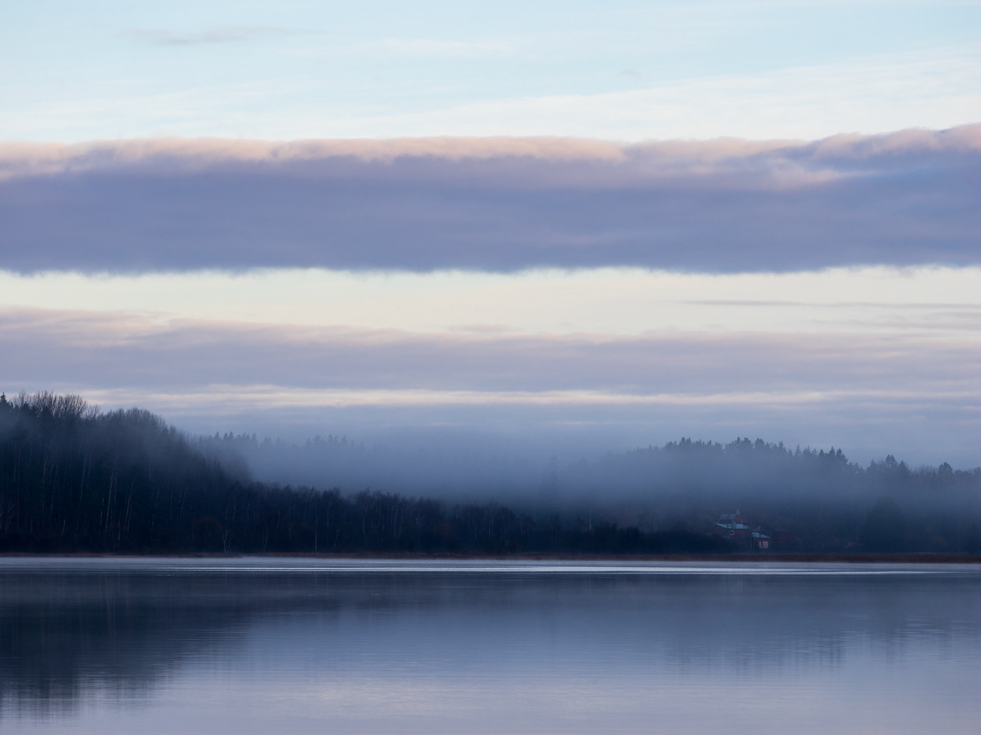 A tranquil lakeside scene shrouded in morning mist, with calm waters reflecting the overcast sky and surrounding forest.