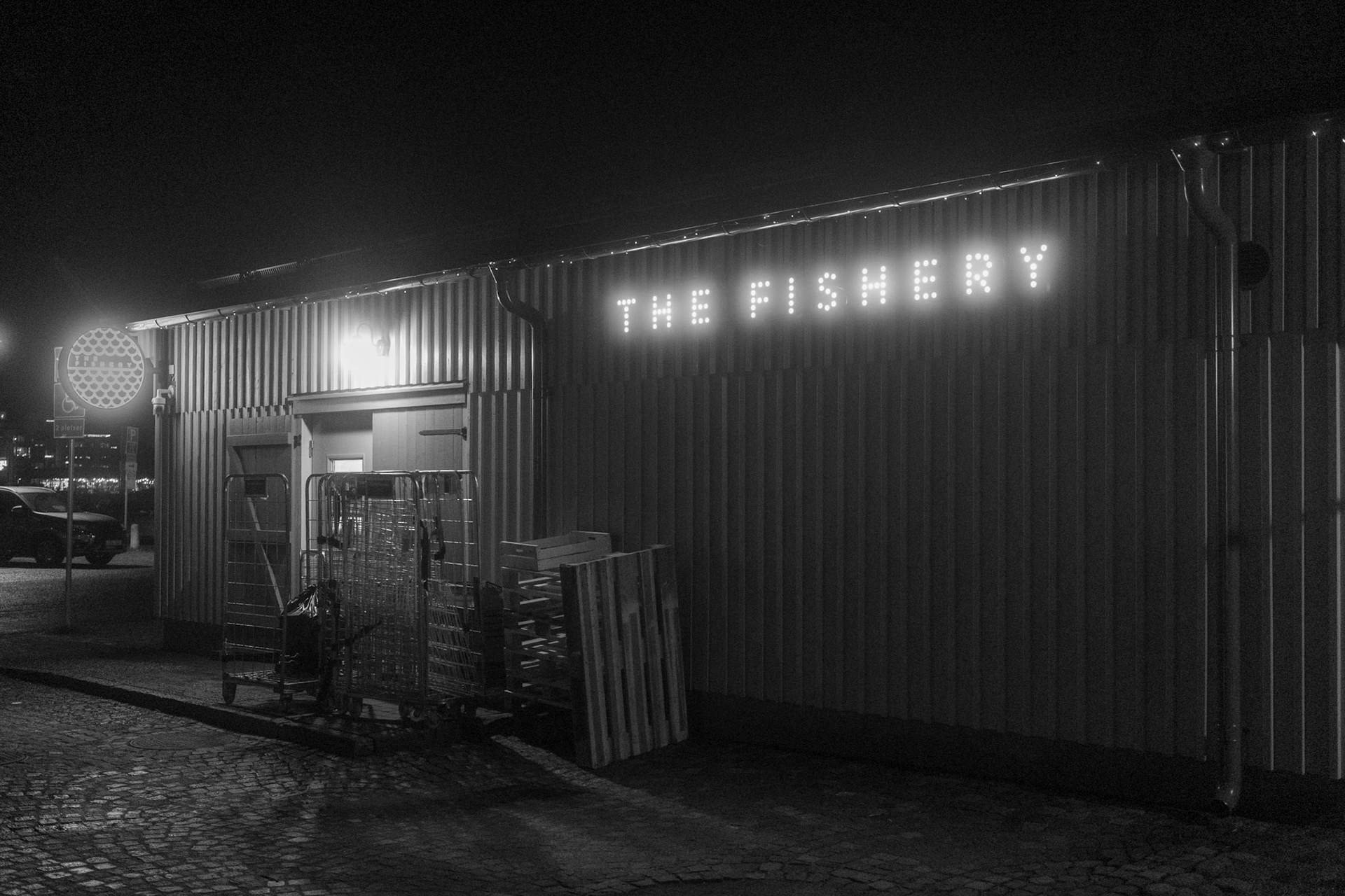 A nighttime scene featuring a modern restaurant housed in a repurposed shipping container. The establishment is illuminated with neon lights, casting a warm glow on the urban street.