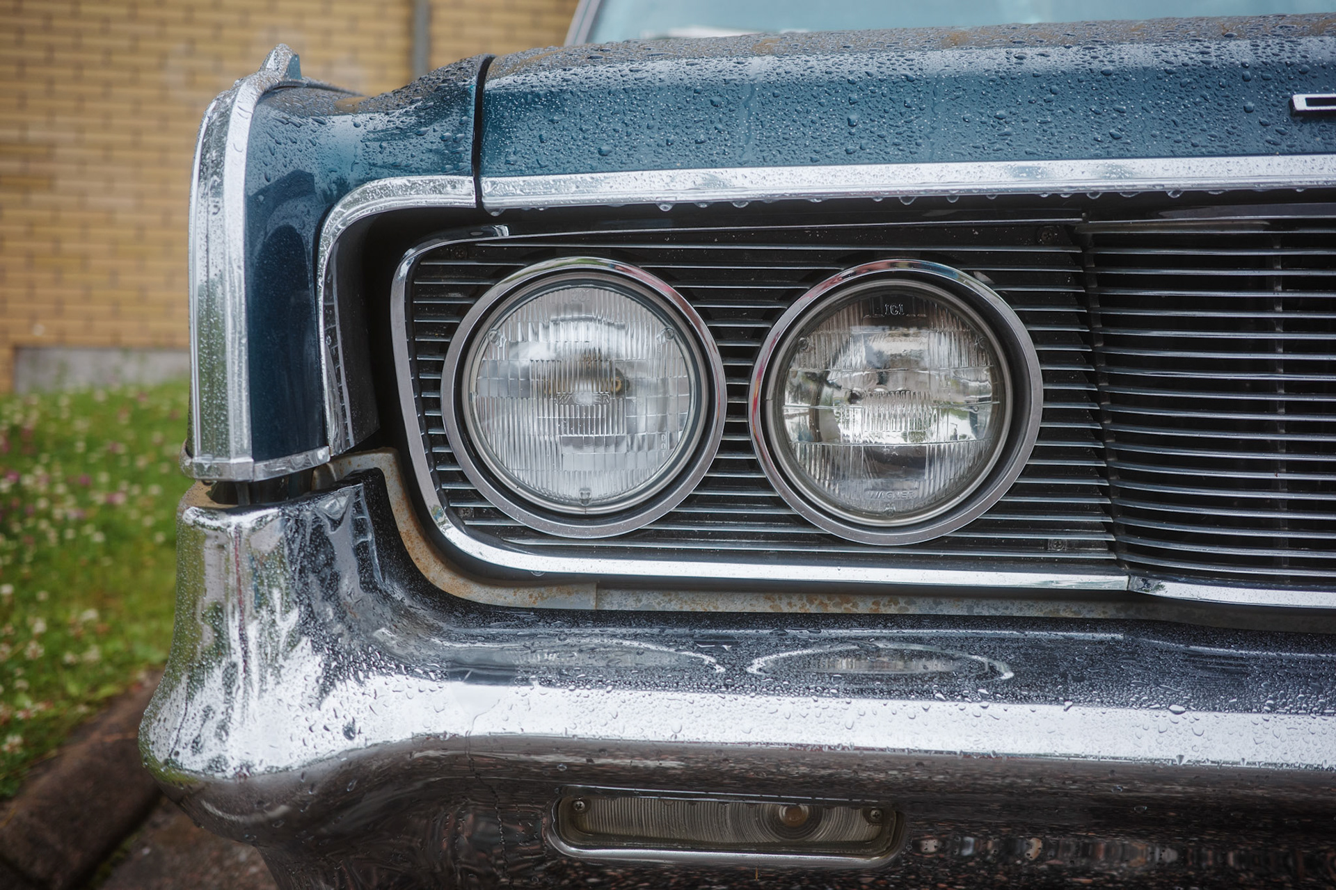A close-up view of a classic car's front end, featuring its distinctive headlights and chrome grill, after a rain shower.