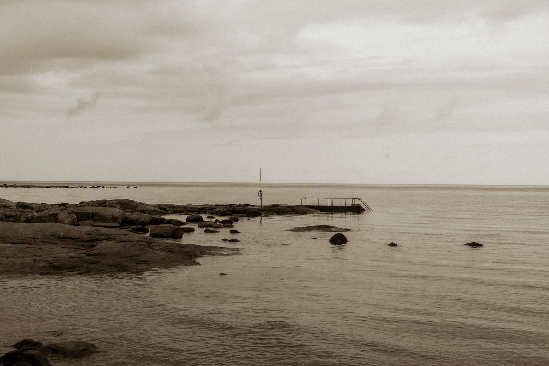 A solitary figure stands on a pier extending into the sea, surrounded by a rocky shoreline under a cloudy sky.