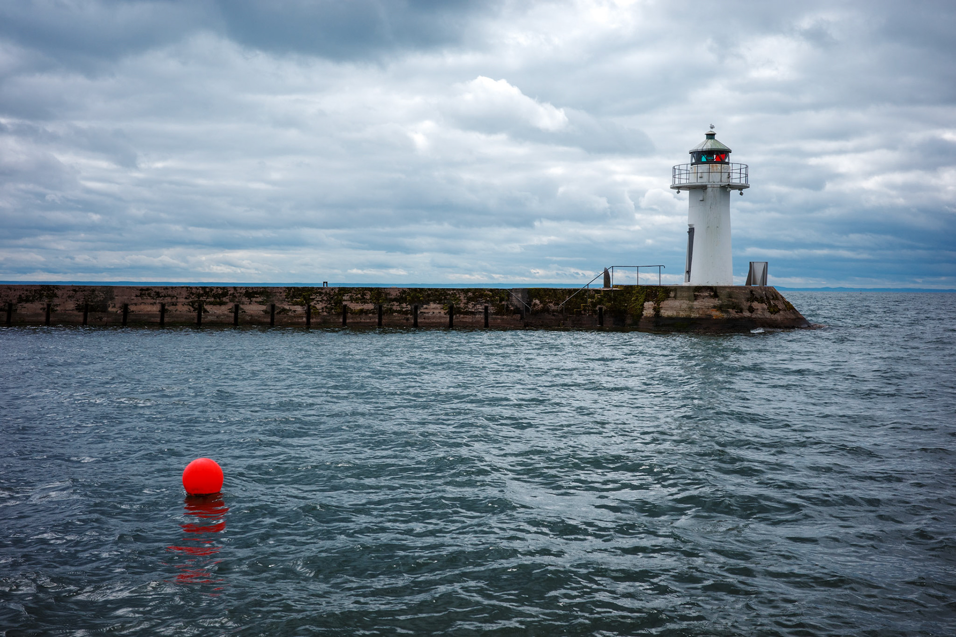 A solitary lighthouse stands at the end of a pier on a cloudy, stormy day, with a red buoy floating in the choppy waters nearby.