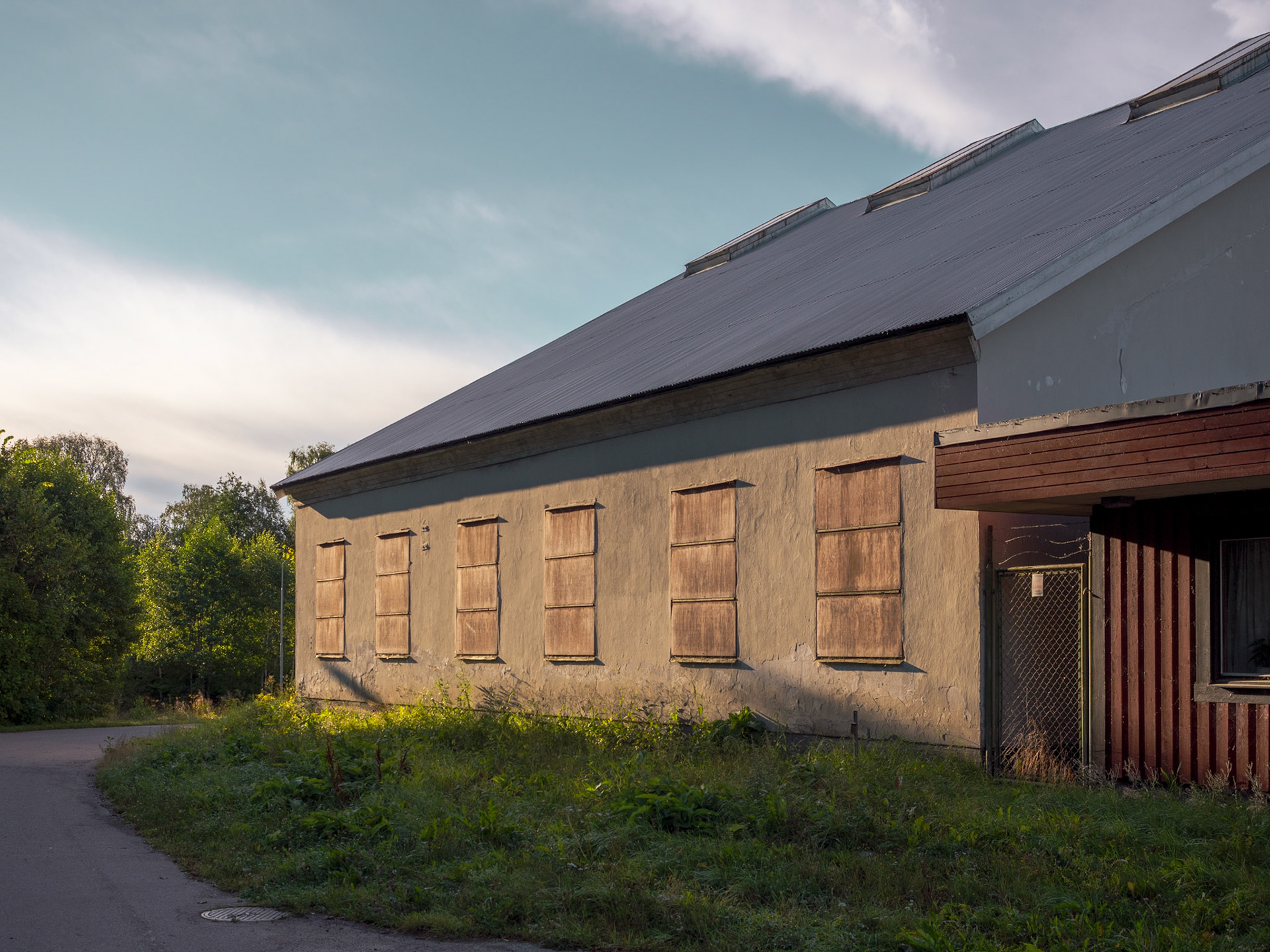 An old, abandoned building with boarded windows and wooden shutters sits along a grassy path, surrounded by overgrown vegetation.