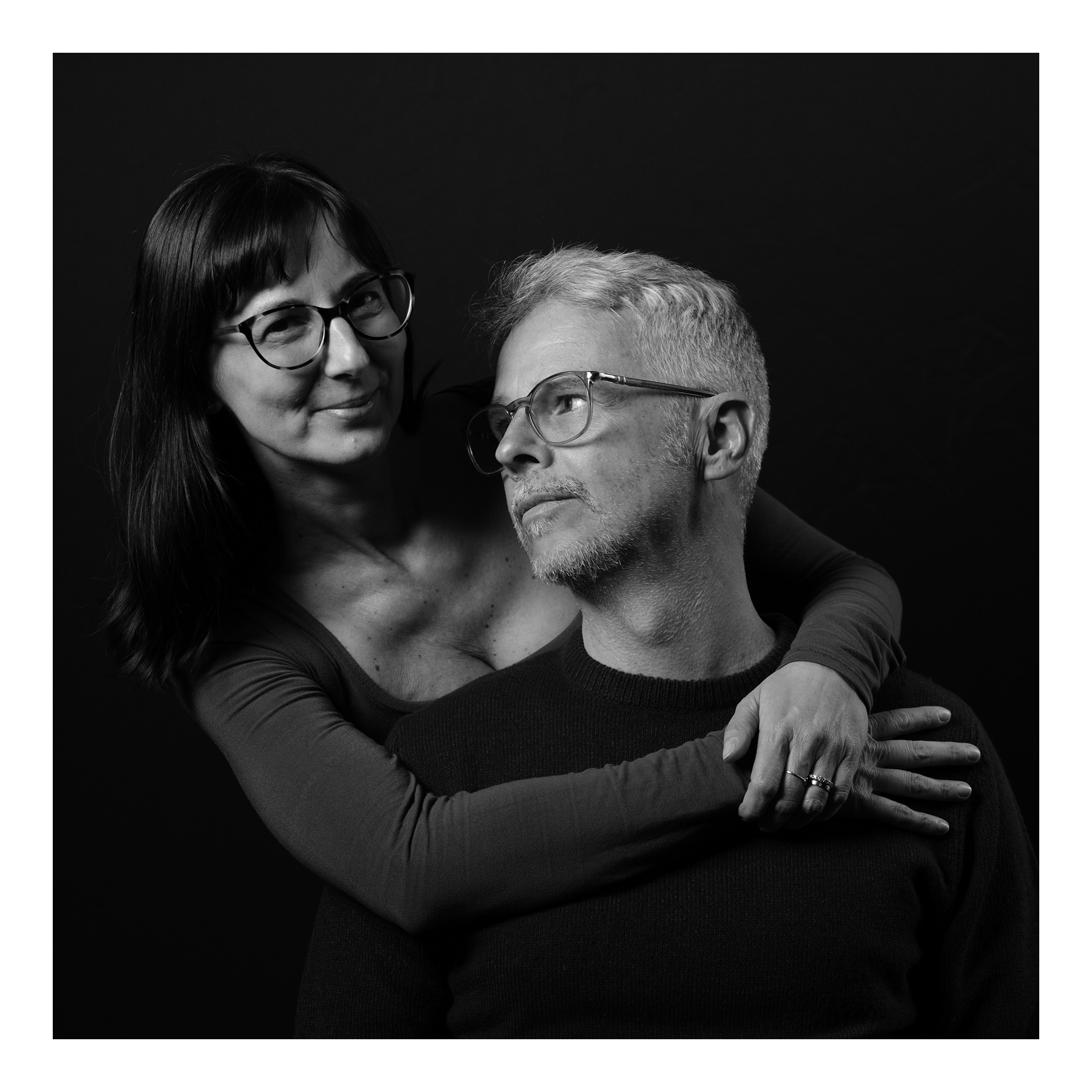 Ritratto fotografico in studio di una coppia con lei che lo abbraccia da dietro mentre lui la guarda