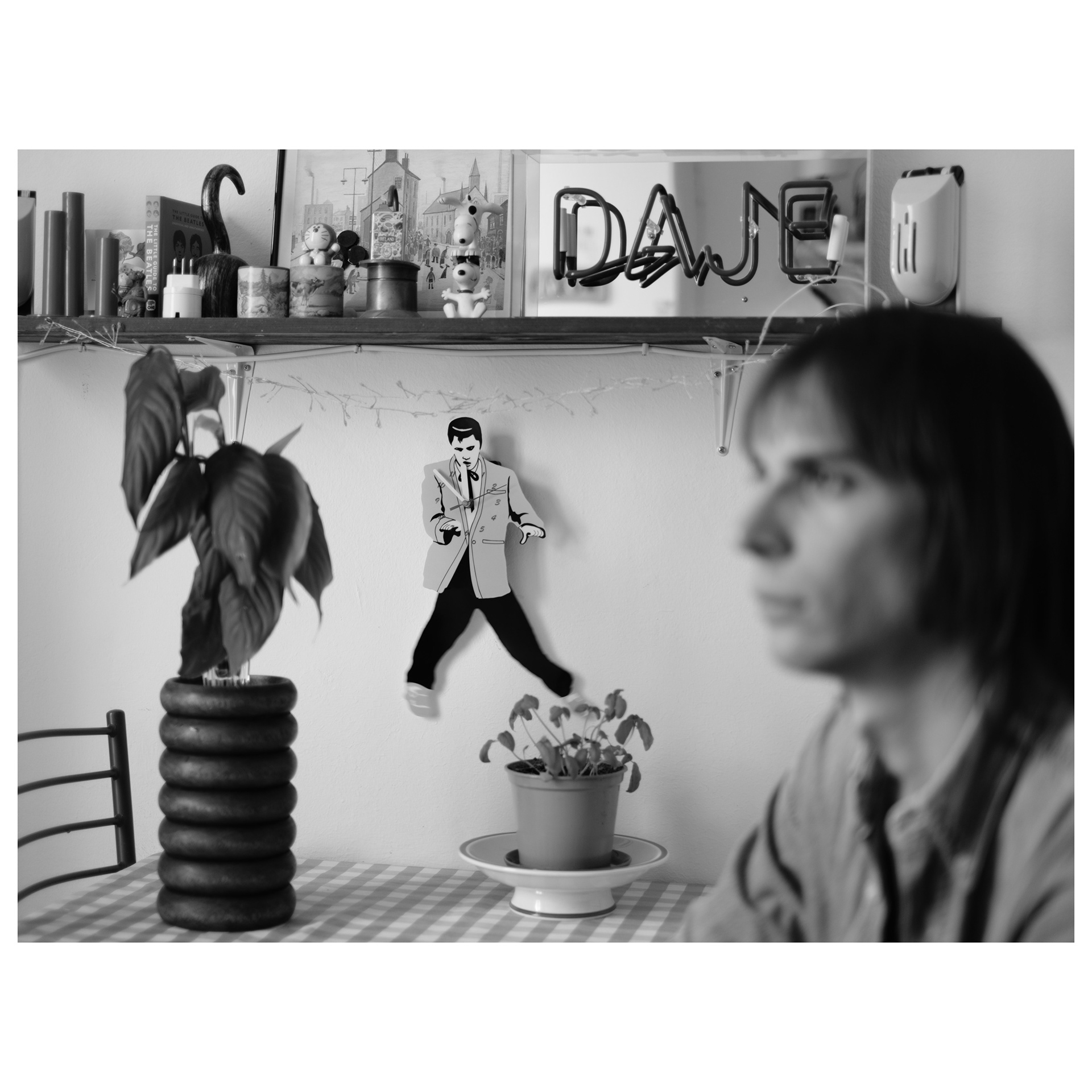 Foto in bianco e nero con in primo piano Alessio sfocato e in secondo piano un tavolo, una pianta e un orologio da parete di Elvis Presley - progetto fotografico di Andrea Palumbo
