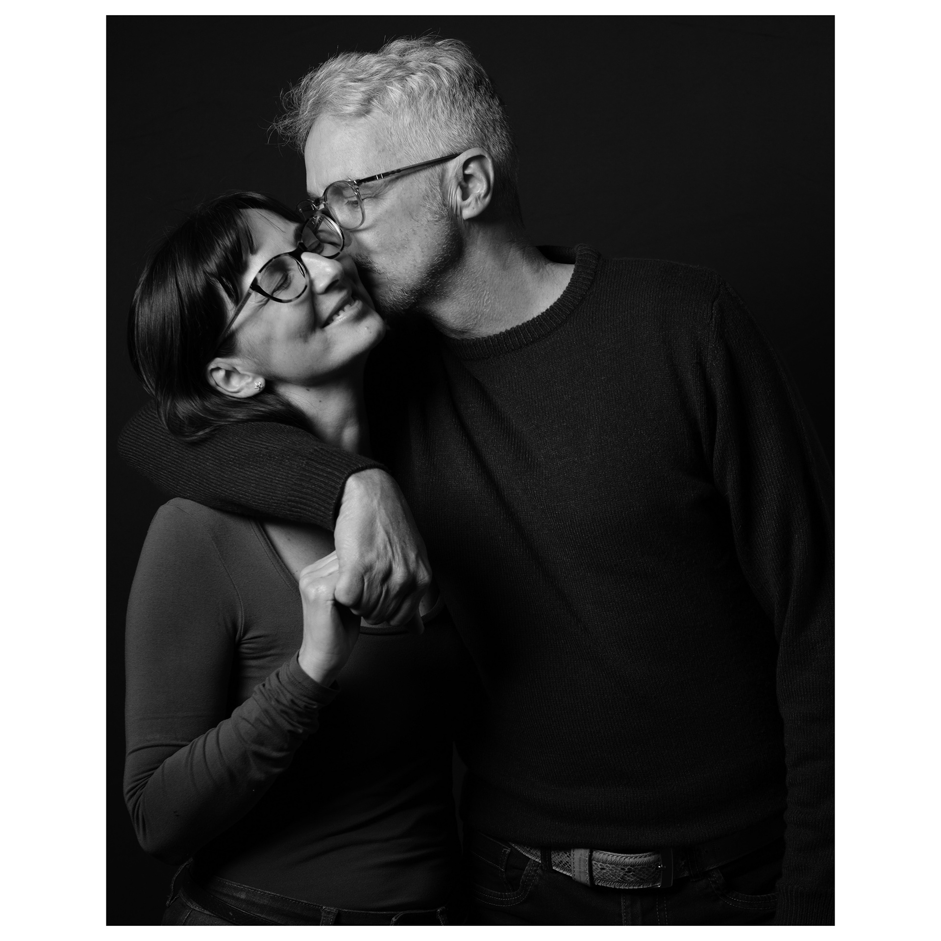 Ritratto fotografico in studio di una coppia abbracciata con lui che la bacia sulla guancia 