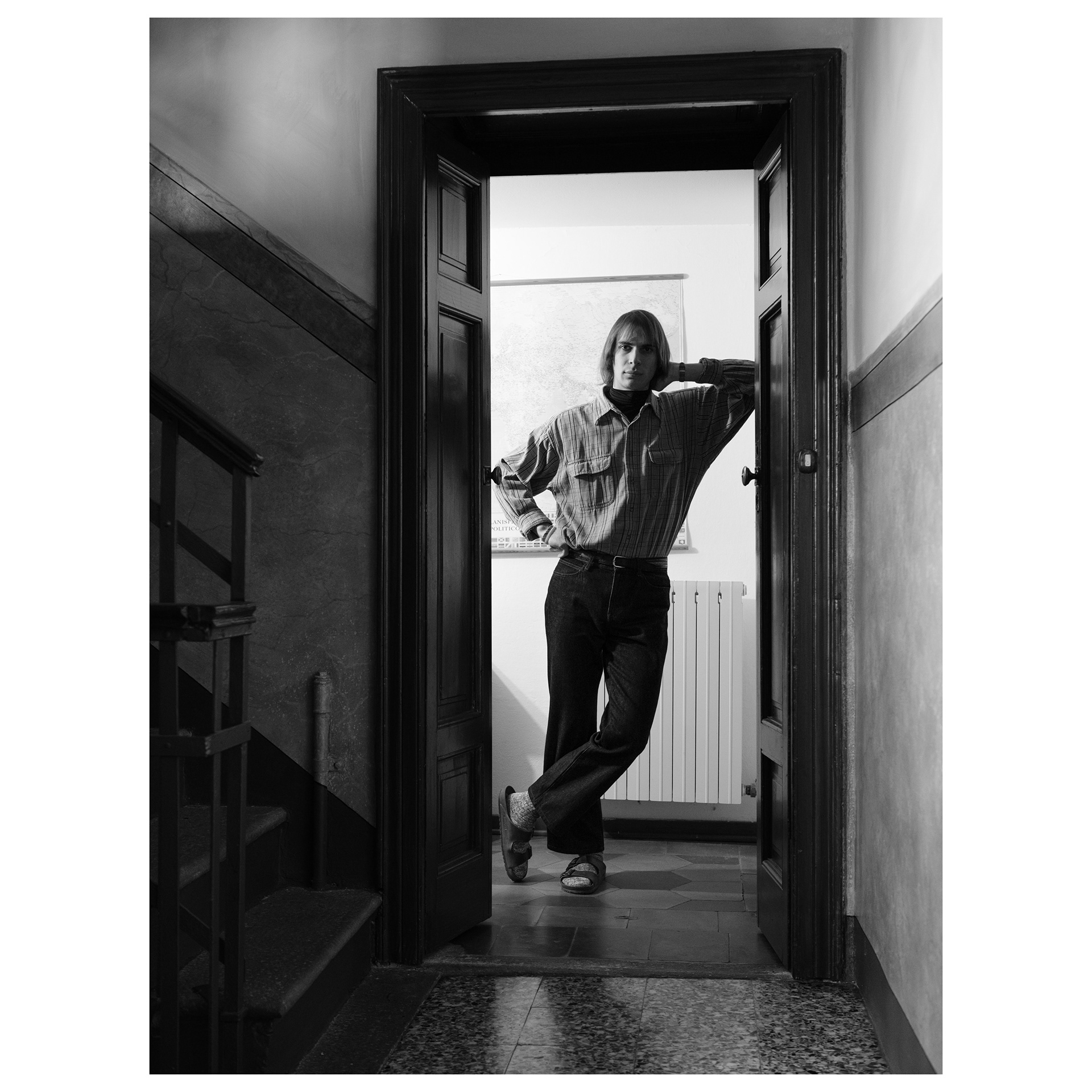 Ritratto in bianco e nero in luce naturale di Alessio appoggiato alla porta di ingresso spalancata del suo appartamento - progetto fotografico di Andrea Palumbo