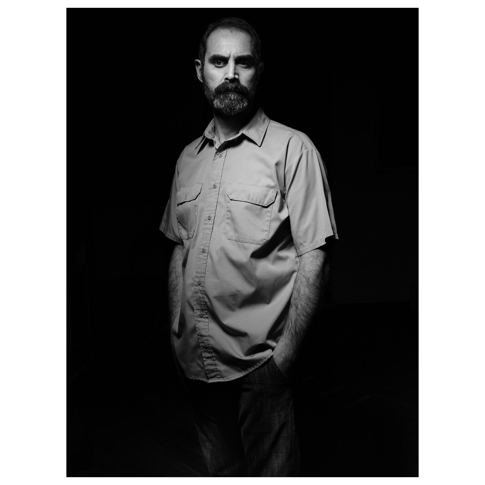 Ritratto in studio in bianco e nero di un uomo con barba dal viso severo e immerso nel buio