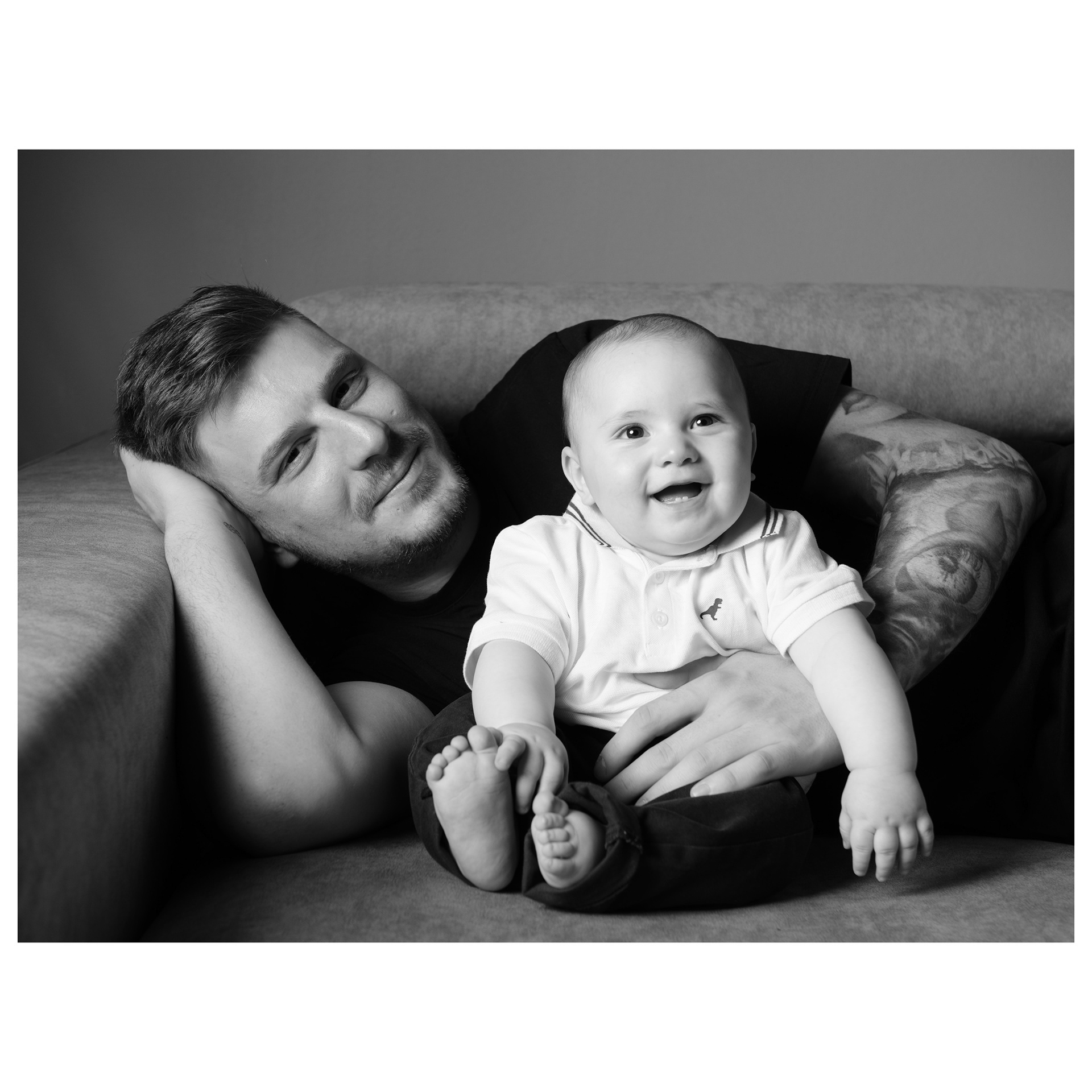 ritratto fotografico in bianco e nero di un papà sorridente e giovane sdraiato sul divano che abbraccia suo figlio molto piccolo avvolti da una luce morbida - Andrea Palumbo fotografo