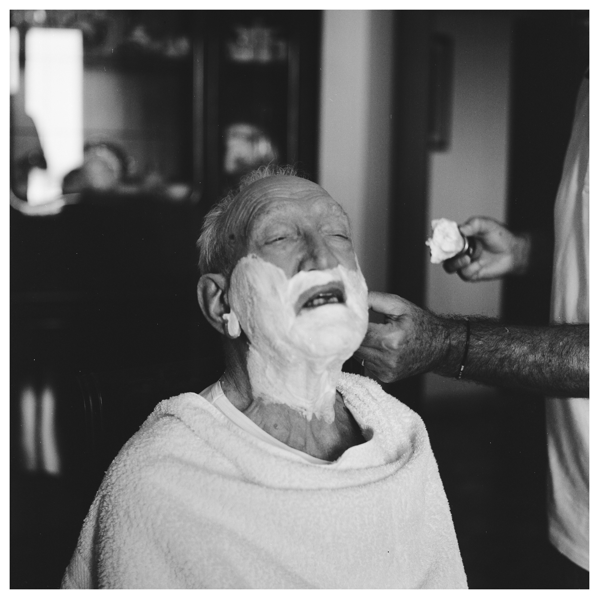 Ritratto in bianco e nero in luce naturale di anziano che si fa rasare la barba in casa dal figlio - progetto autoriale Andrea Palumbo fotografo