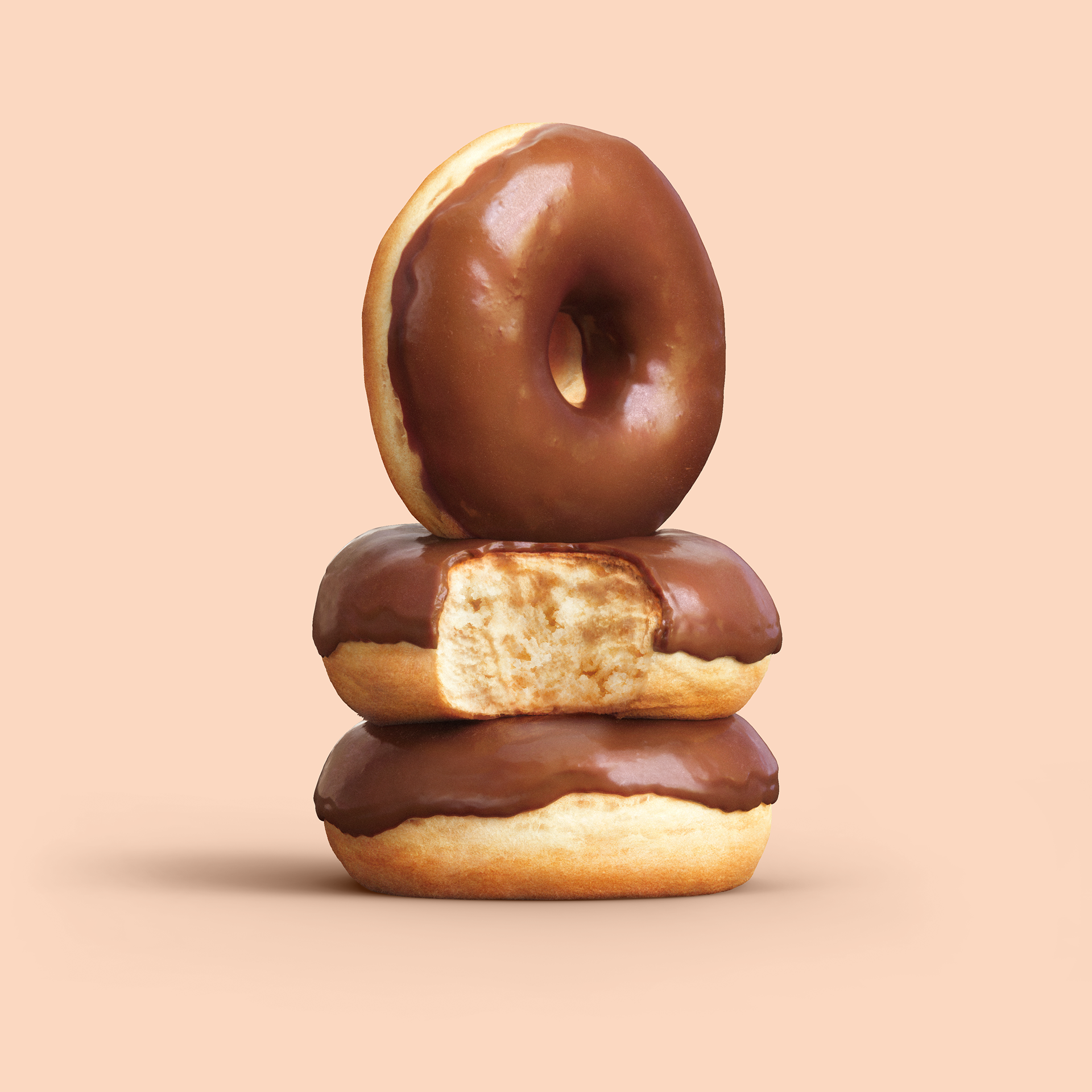Three chocolate-glazed doughnuts stacked on top of each other, with one partially eaten, against a pastel background.