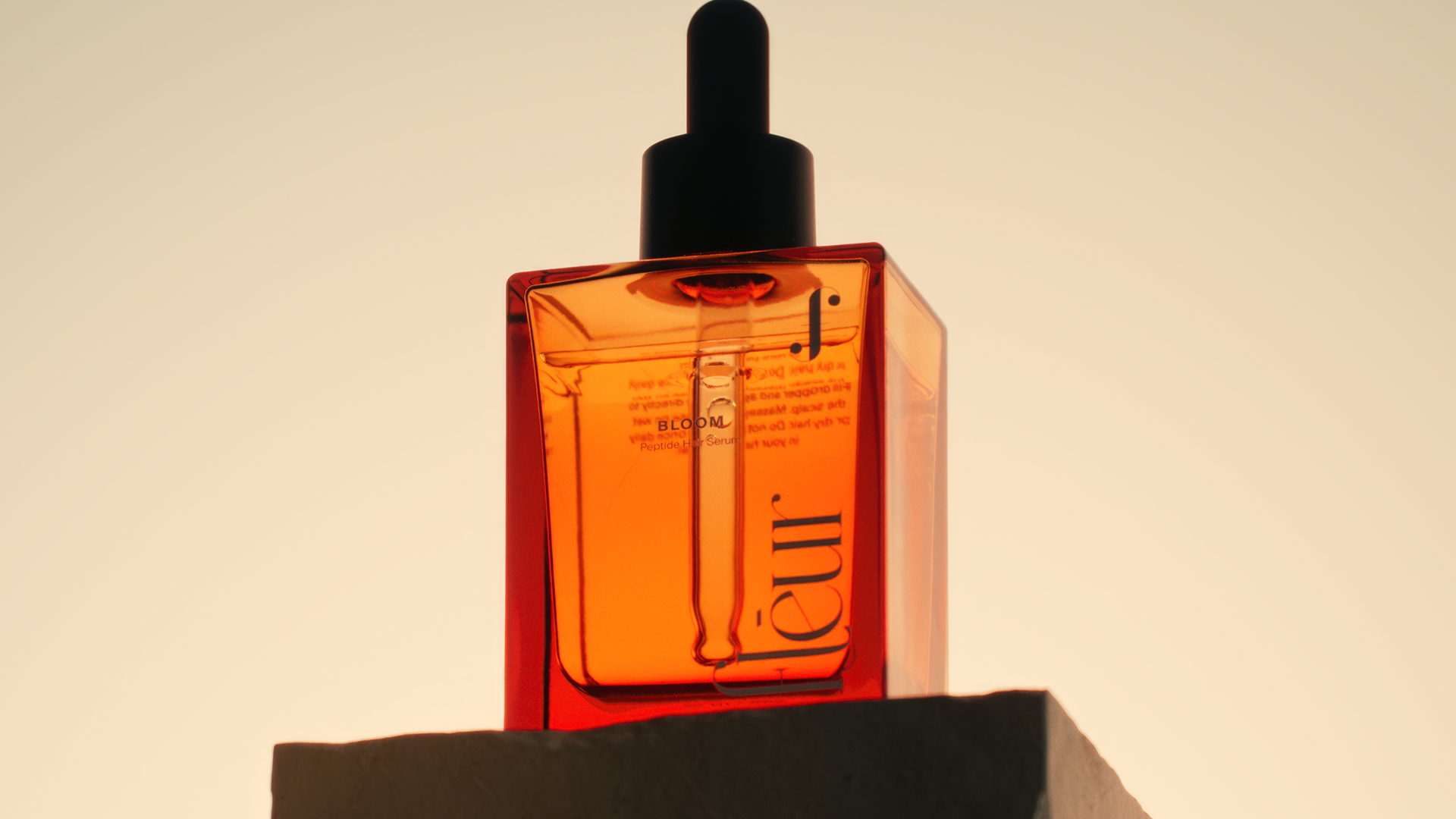A sleek, amber-colored glass bottle with a dropper cap, featuring the brand name "J." and product name "BLOOM" on its side.