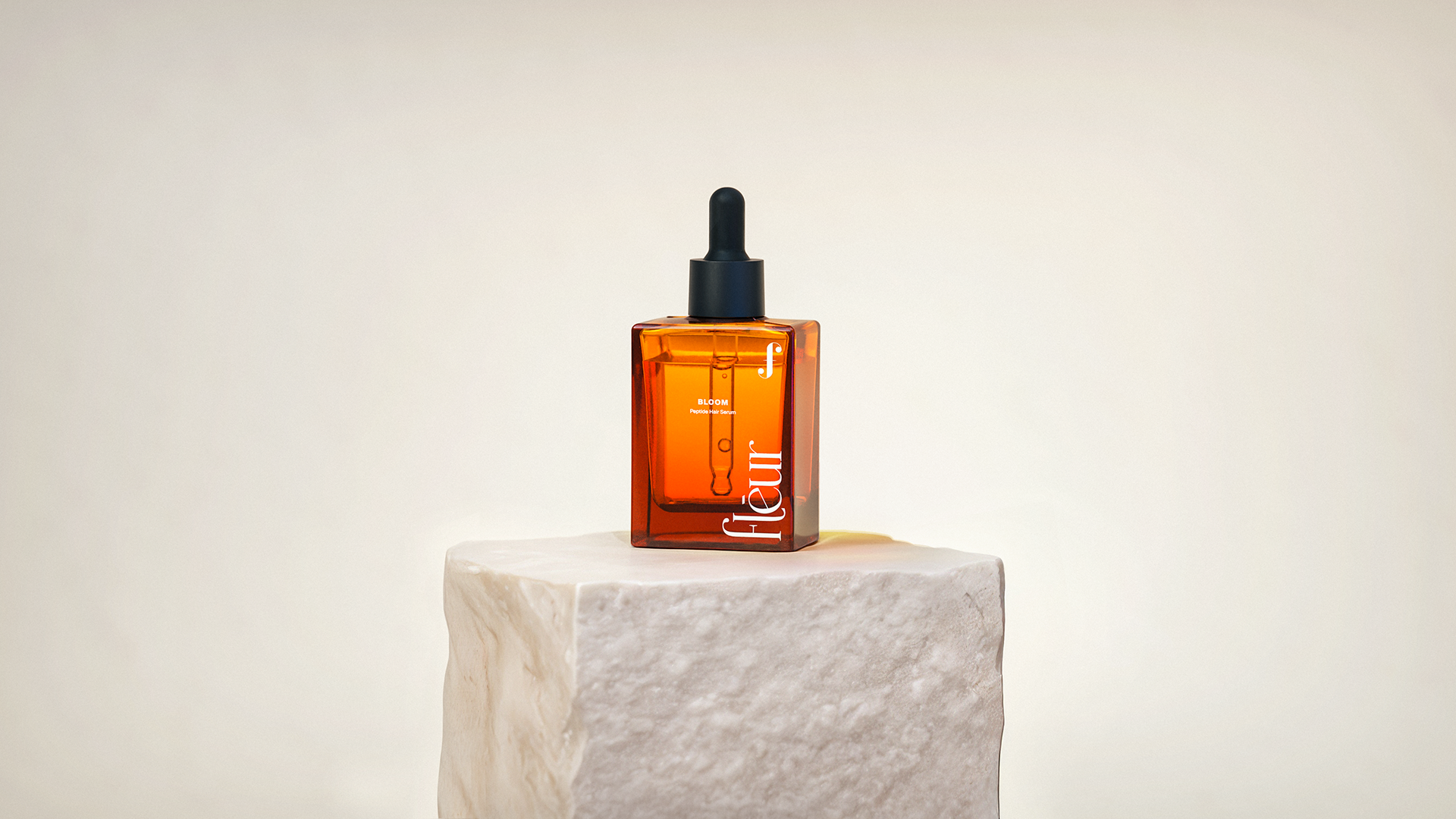 A sleek, amber-colored bottle of "Fleur" hair serum stands on a white pedestal against a neutral background.