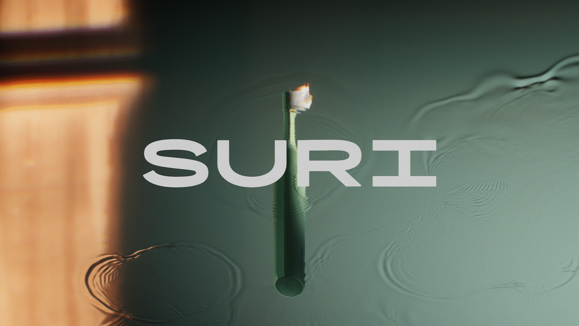 Animated sequence: A green, elongated object resembling a toothbrush is partially submerged in water, with ripples spreading outwards. The word "SURI" is prominently displayed in bold, white letters across the center of the image.