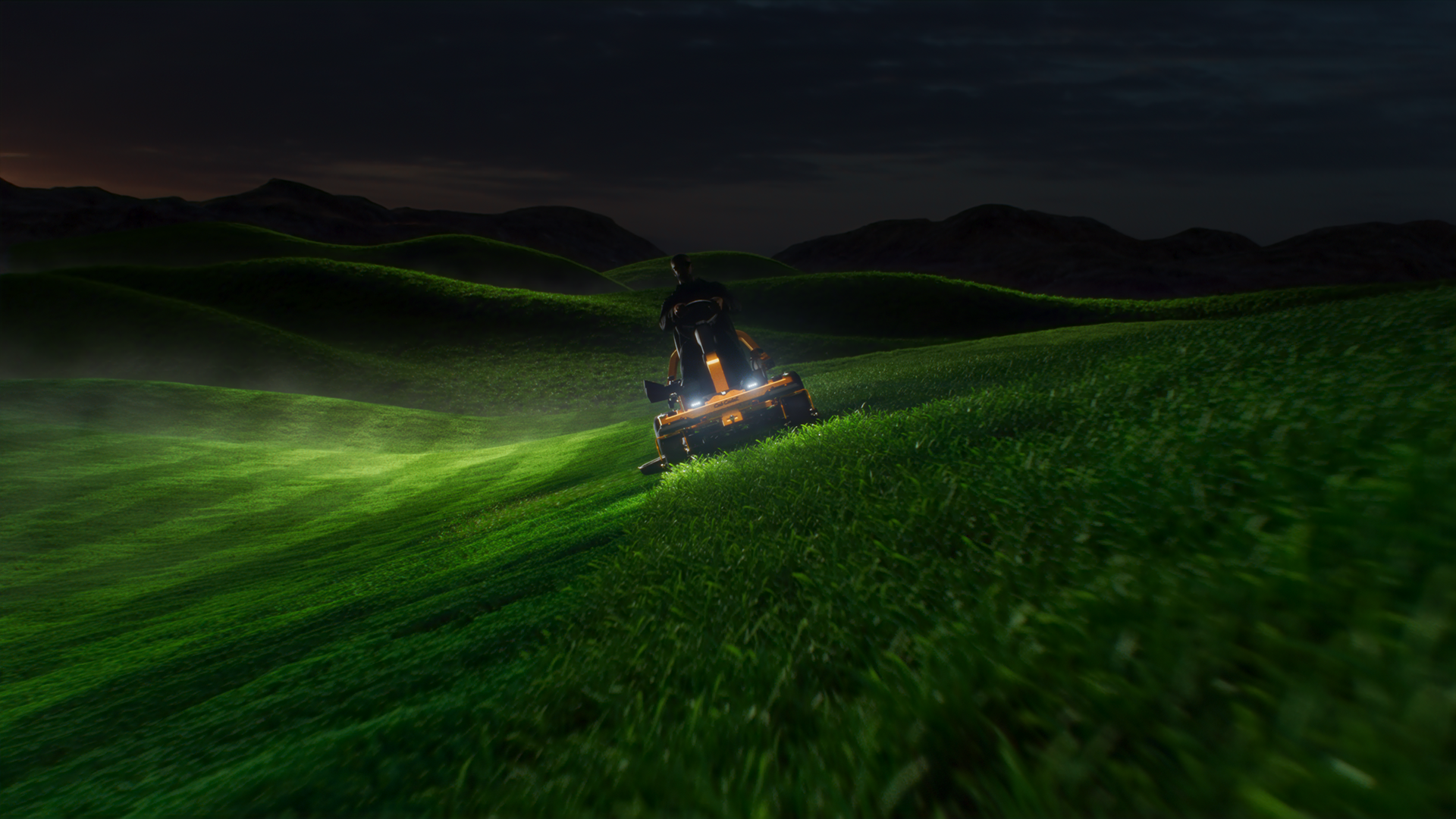 A lawn mower with headlights on, cutting grass at night, with mountains in the background.