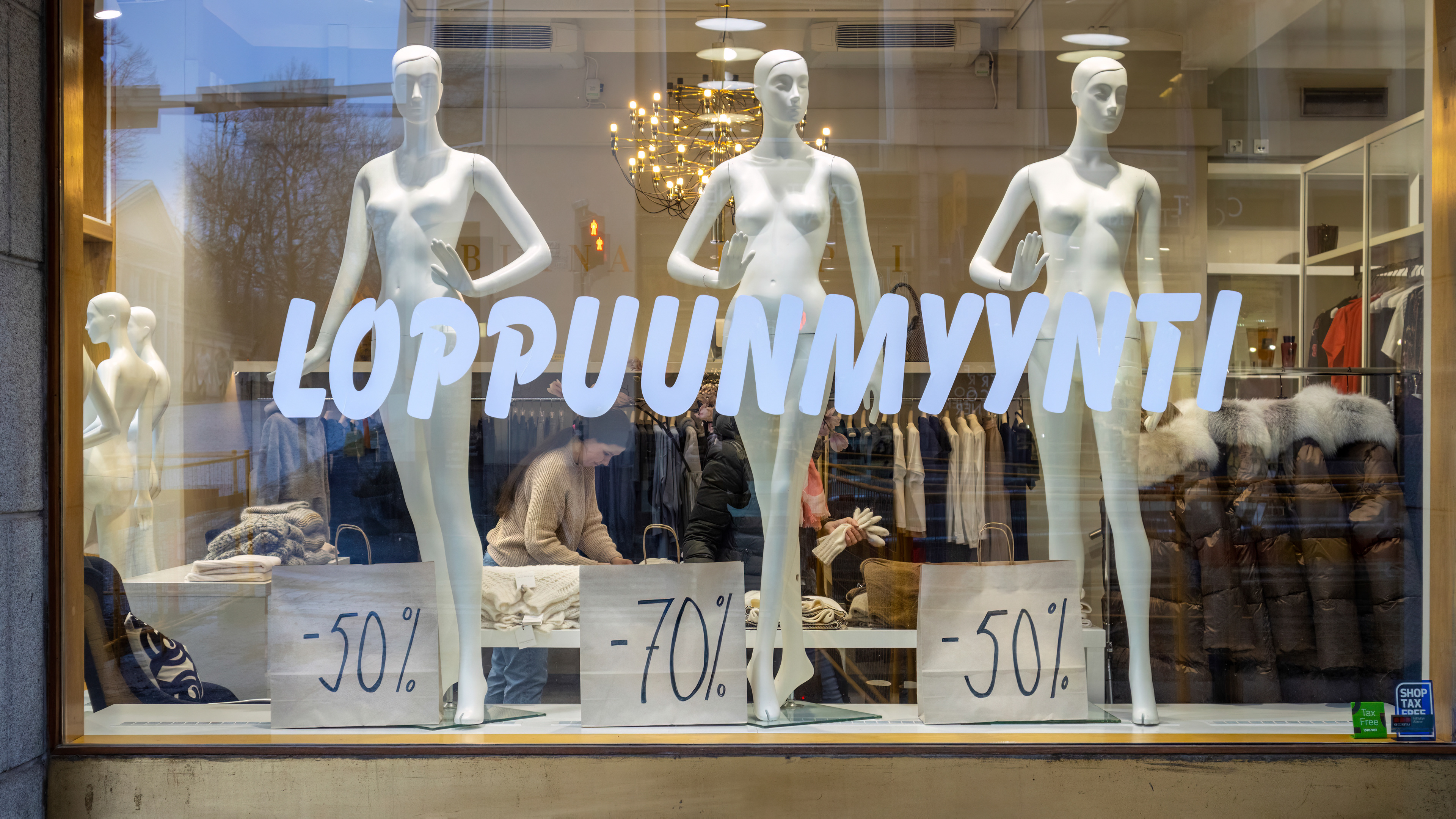 Vaatekaupan näyteikkunassa kolme valkoista mallinukkea ja suuri teksti 'LOPPUUNMYYNTI'. Etualalla paperikyltit, joissa alennusprosentit -50 %, -70 % ja -50 %. Ikkunan takana sisällä näkyy myyjä ja asiakas sekä talvivaatteita kirkkaasti valaistussa liikkeessä.