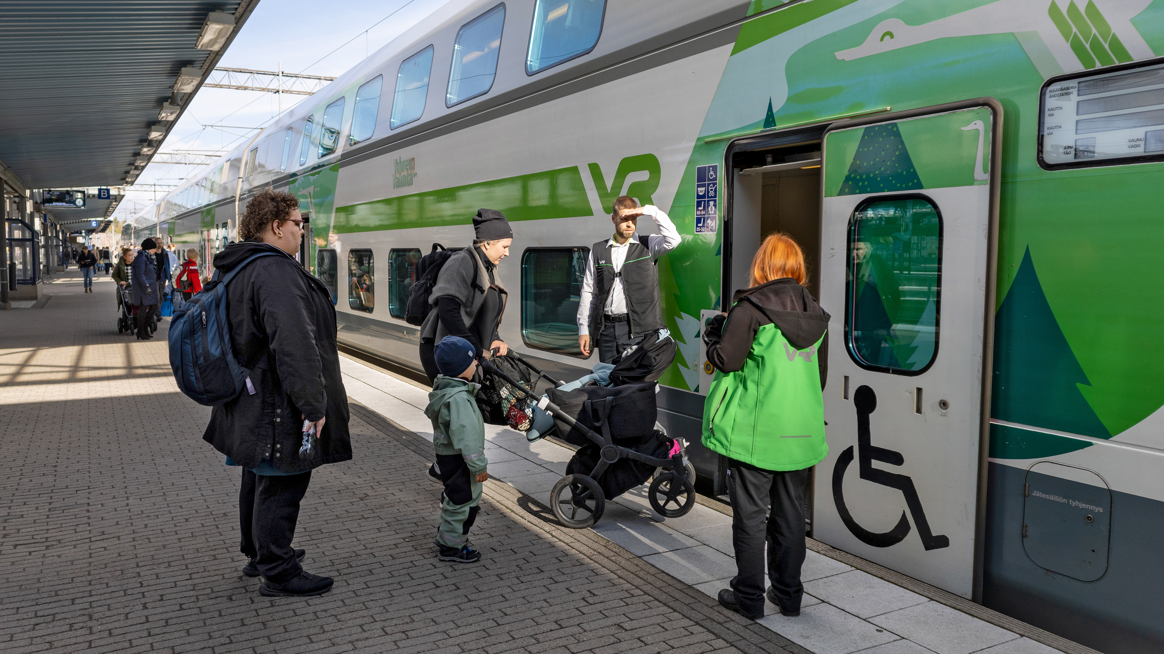 VR:n vihreäsävyinen kaksikerroksinen juna on pysähtynyt Pasilan asemalle. Junahenkilökunta auttaa äitiä, jolla on lastenvaunut ja pieni lapsi, nousemaan junaan. Vieressä seisoo useita matkustajia, ja taustalla näkyy lisää ihmisiä asemalaiturilla auringonpaisteessa.
