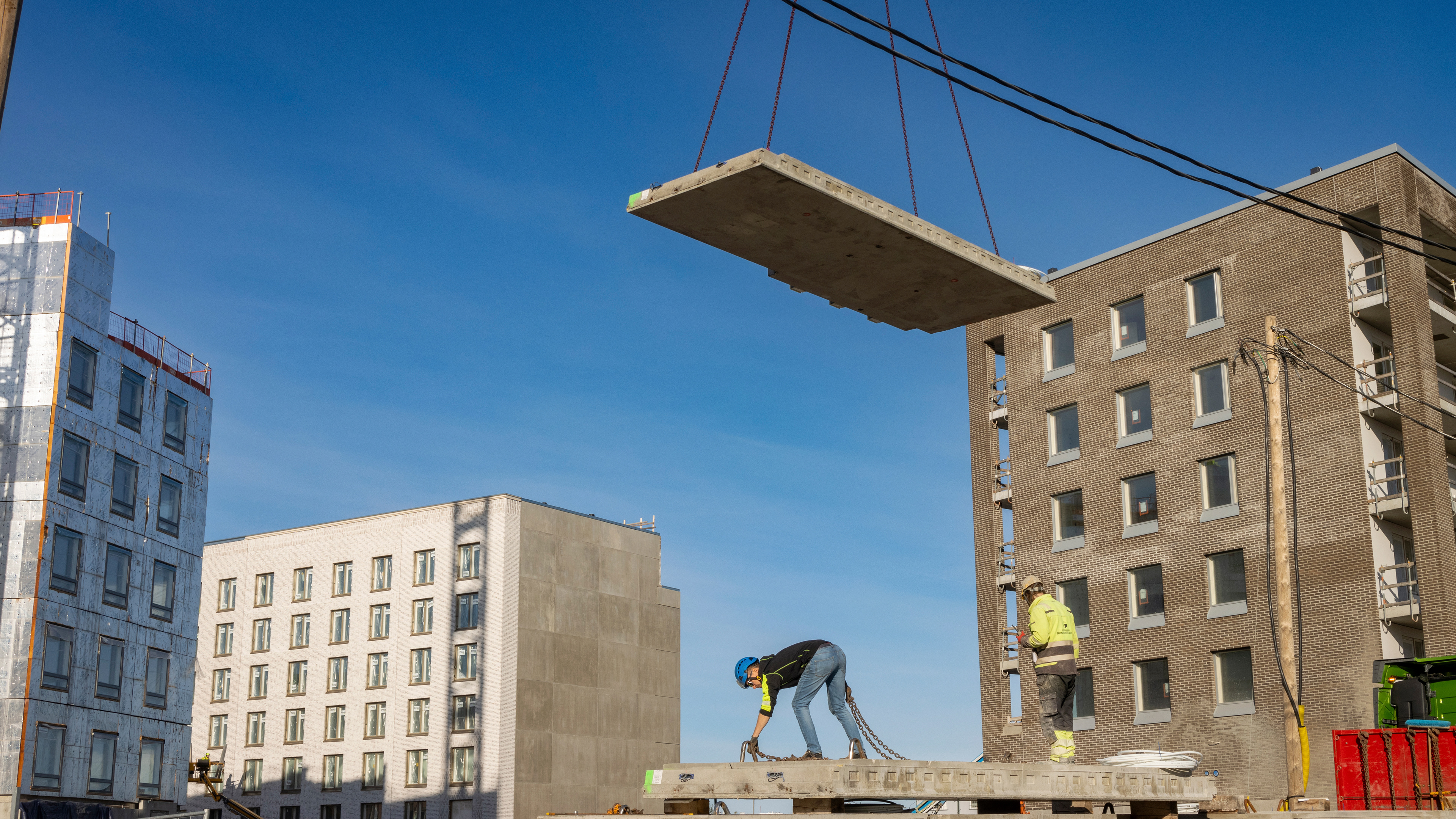 Kaksi rakennustyöntekijää työskentelee kerrostalotyömaalla Verkkosaaressa. Toinen kiinnittää ketjuja betonilaattaan, toinen seisoo vieressä. Yläpuolella nosturi laskee suurta betonilevyä paikoilleen kirkkaan sinisen taivaan alla.