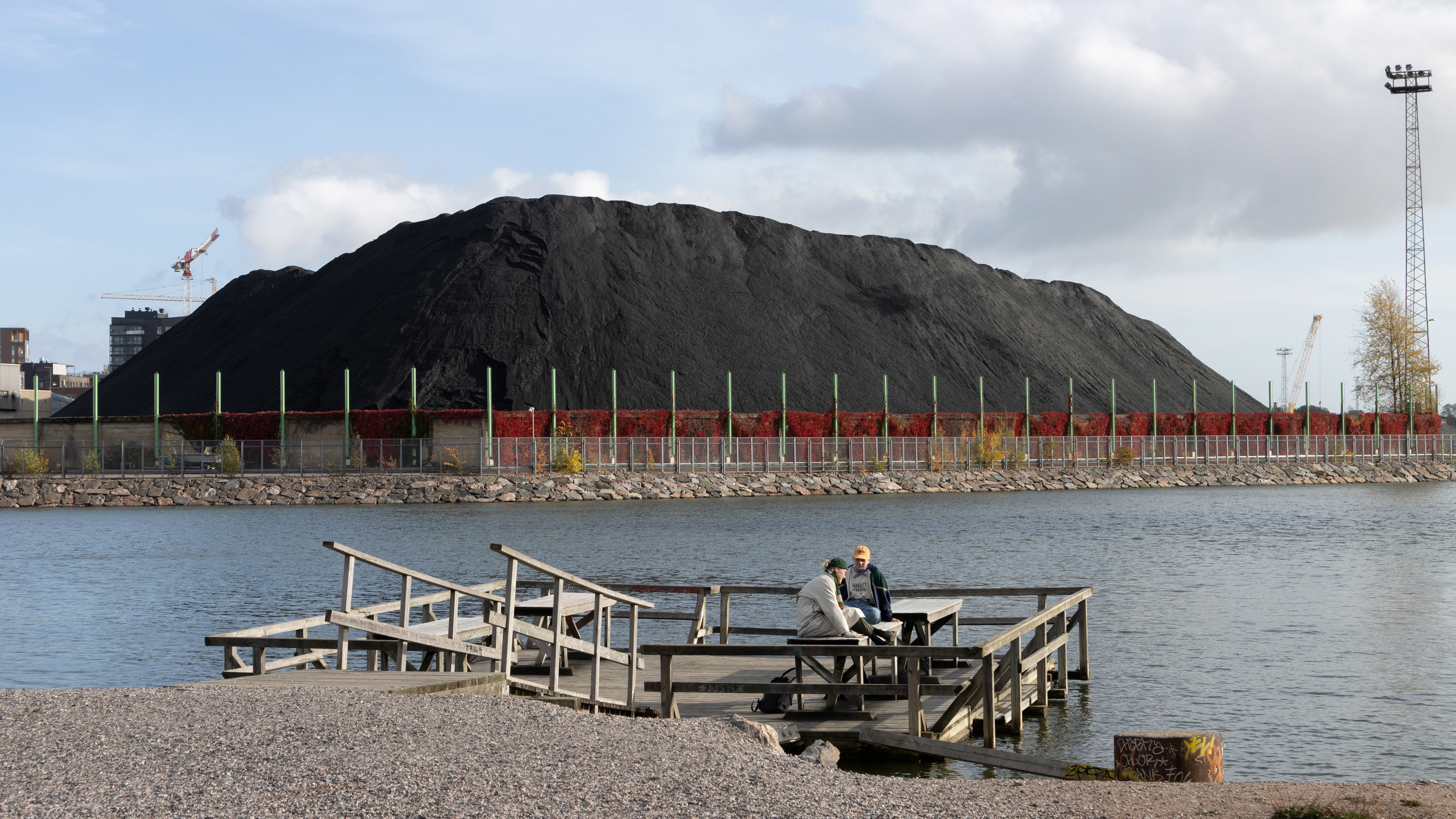 Kaksi ihmistä istuu puisella mattolaiturilla veden äärellä keskustelemassa. Taustalla Hanasaaressa näkyy suuri musta kivihiilikasa aidan ja punalehtisten köynnöskasvien takana. Kuvan reunoilla vasten taivasta näkyy nostureita ja kerrostaloja.