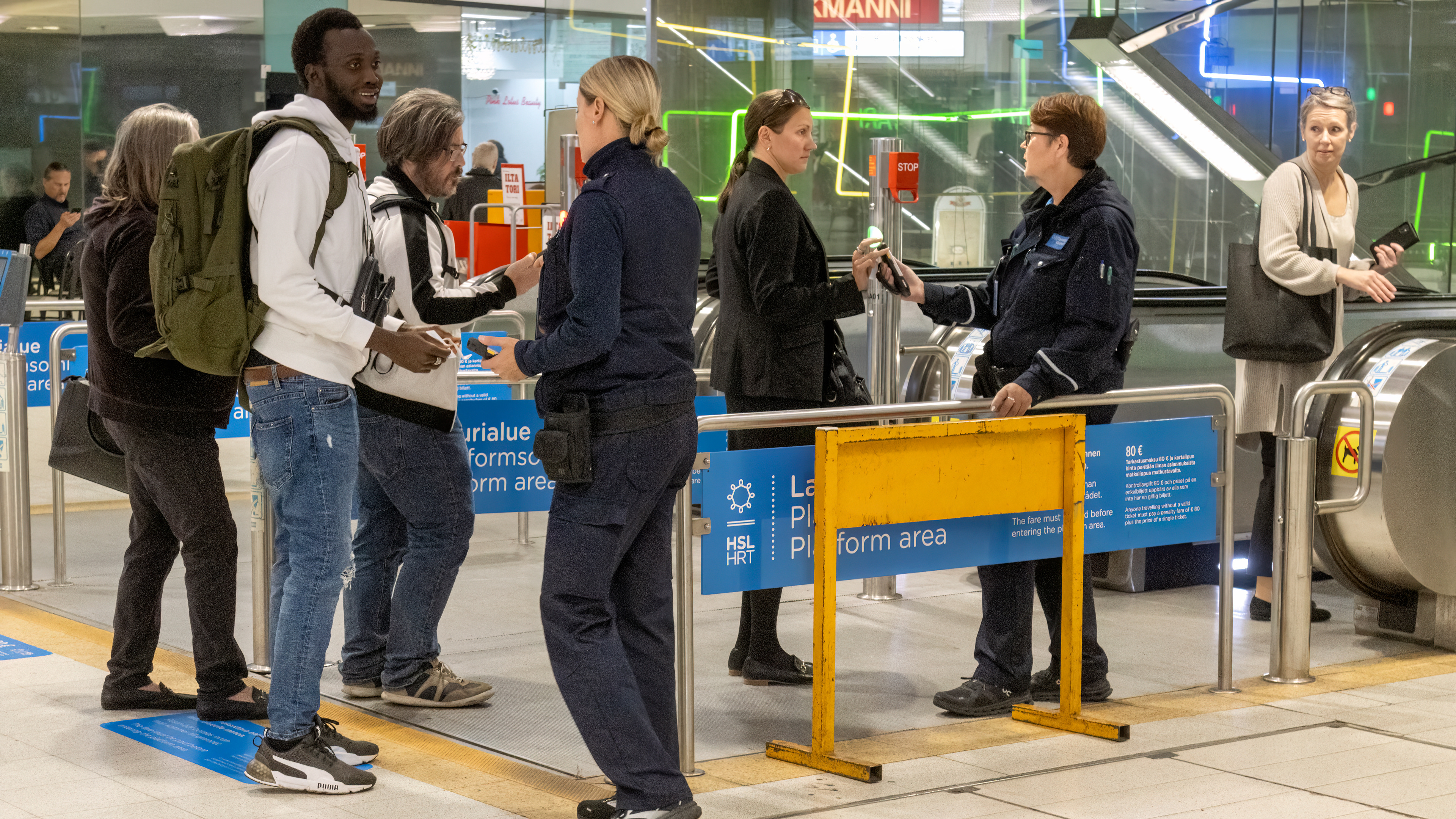 Metron lippuporttien edessä valvojat tarkistavat metroon menevien matkustajien lippuja. Ihmiset jonottavat portille, osa näyttää lippua puhelimesta tai kortilta. Taustalla näkyy liukuportaat ja neonvaloja, sekä HSL:n kyltit, joissa kerrotaan lipun tarkistuksesta ja sakon määrästä.