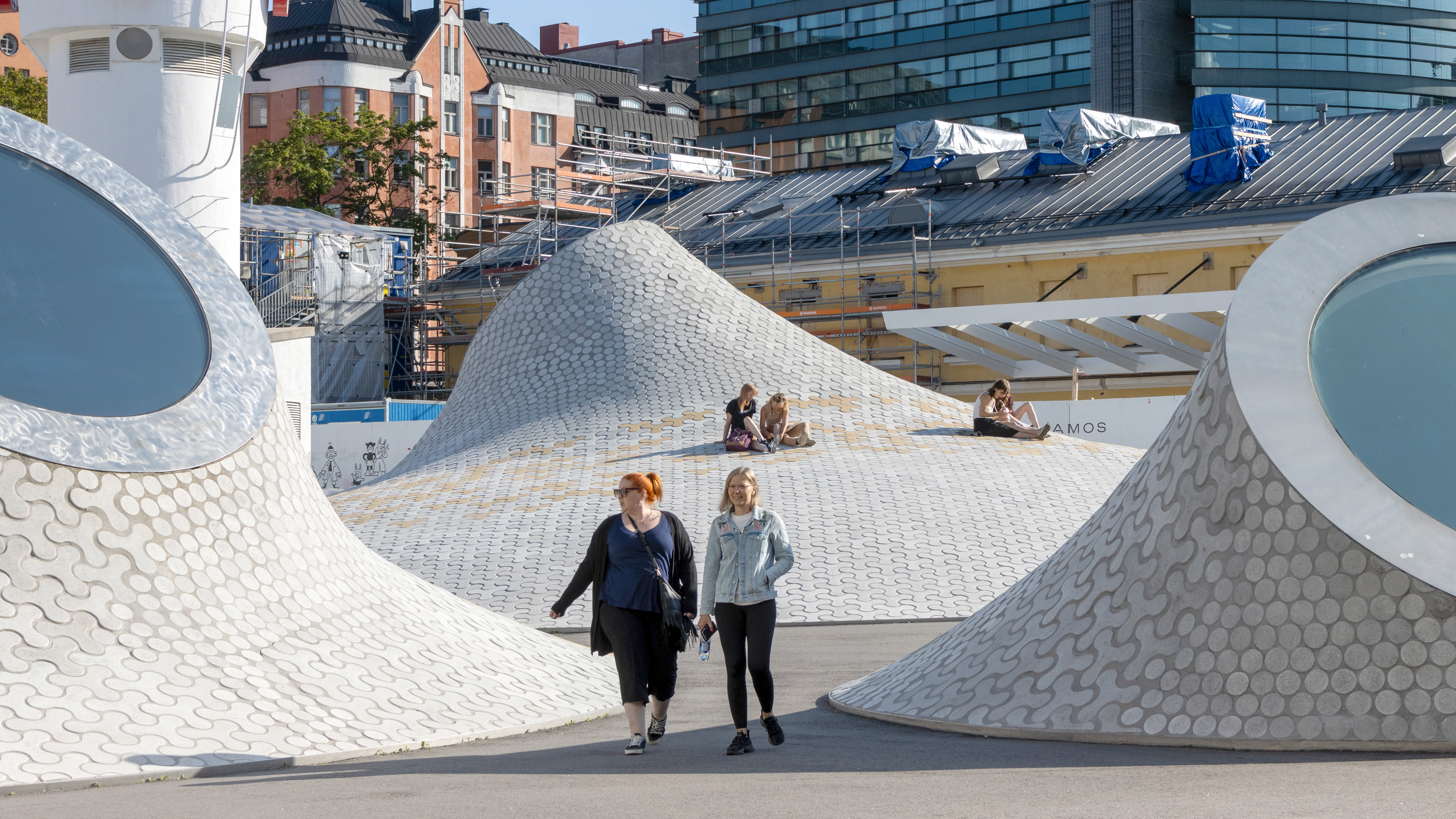 Kaksi naista kävelee kohti Lasipalatsia Helsingissä. Ympärillä näkyy kaarevien kuviollisten pintojen yläosassa Amos Rex -taidemuseon pyöreitä kattoikkunoita. Taustalla ihmiset istuvat ja viettävät aikaa katon kaarevilla pinnoilla. Rakennusten takana näkyy sekä vanhempaa arkkitehtuuria että moderneja lasijulkisivuja.