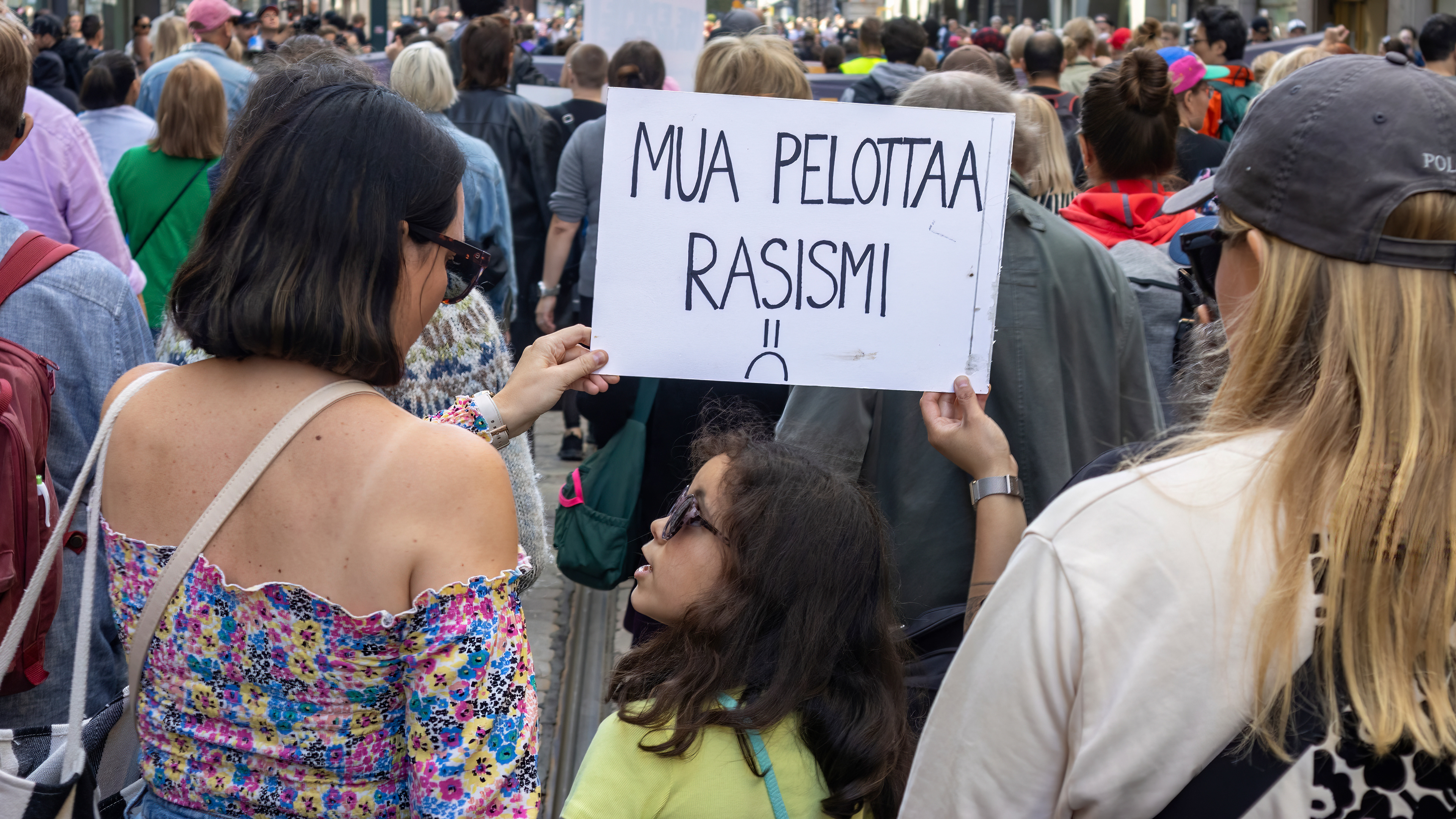 Mielenosoituksessa naiset pitävät käsissään kylttiä, jossa lukee 'Mua pelottaa rasismi'. Keskellä seisoo lapsi aurinkolaseissa ja katsoo toista naisista. Taustalla suuri joukko ihmisiä kävelee kadulla, monilla on kylttejä ja banderolleja.