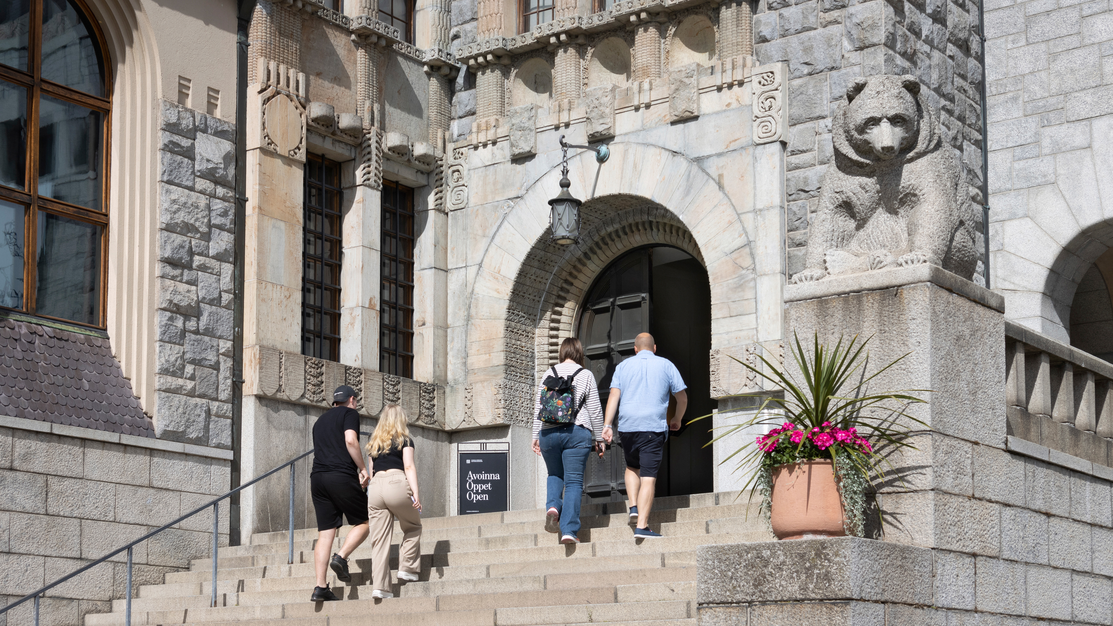 Kaksi pariskuntaa kävelee ylös kivisiä portaita kohti Kansallismuseon sisäänkäyntiä, jonka vieressä on kivinen karhupatsas. Ovi on auki ja sen vieressä on kyltti, jossa lukee 'Avoinna Öppet Open'. Karhupatsaan edessä tasanteella on kukkaruukku, jossa on punaisia kukkia.