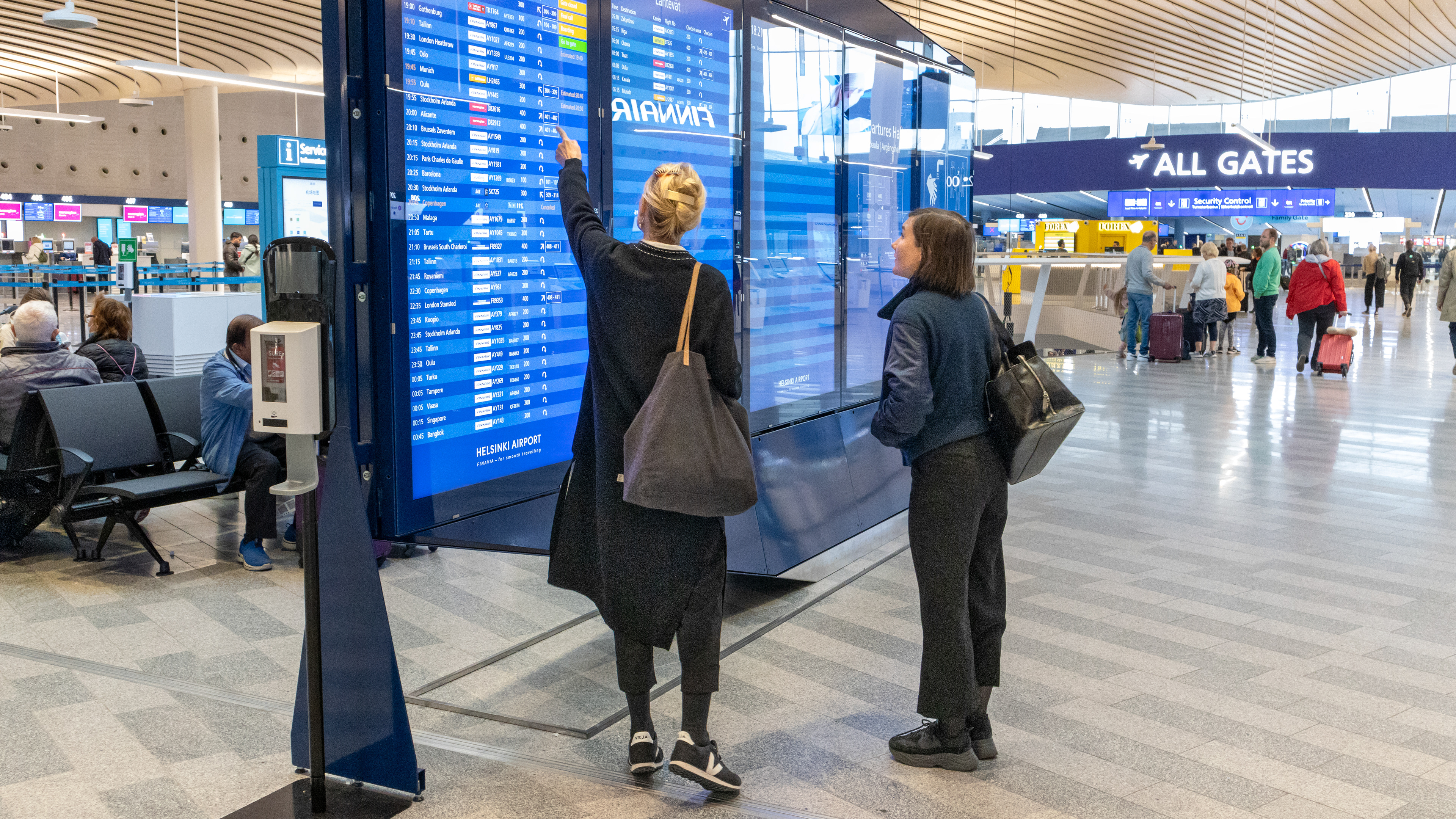 Kaksi naista katsoo ja toinen osoittaa suurta lähtöaikataulunäyttöä Helsinki-Vantaan lentoasemalla. Taustalla näkyy matkustajia, jotka odottavat tai kävelevät kohti turvatarkastusta, sekä opaste 'All Gates'. Hallissa on moderni sisustus ja kaareva, vaalea katto.