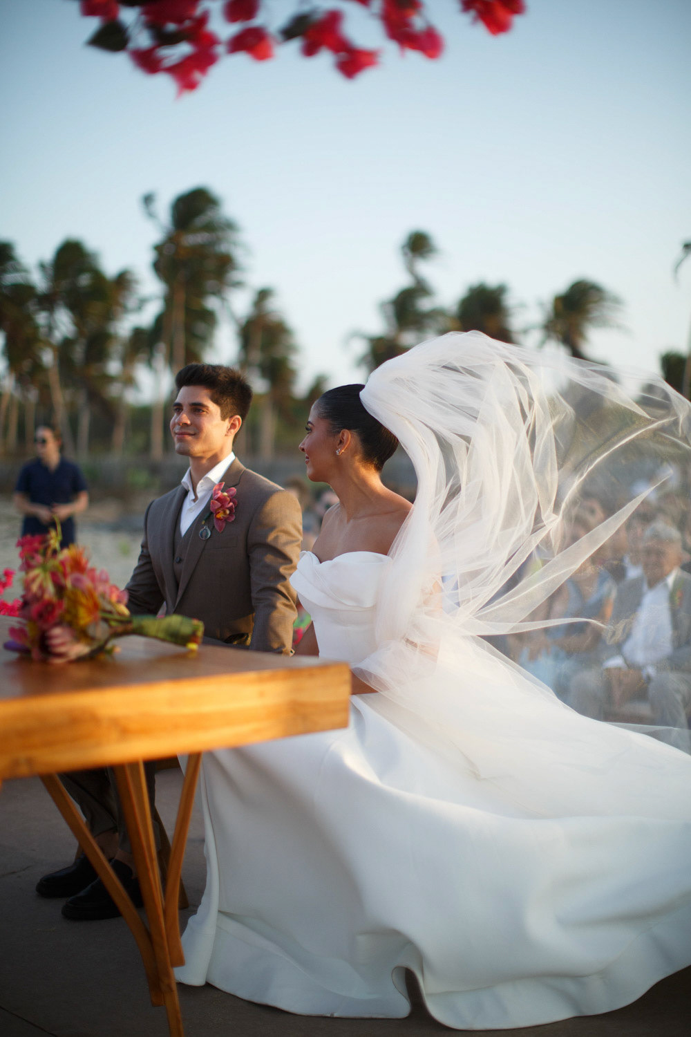 Noiva e noivo no altar durante cerimônia na praia com véu ao vento e terno slim marrom. Fotografia de Rebeca Santos na praia de Barra Grande, litoral do Piauí.