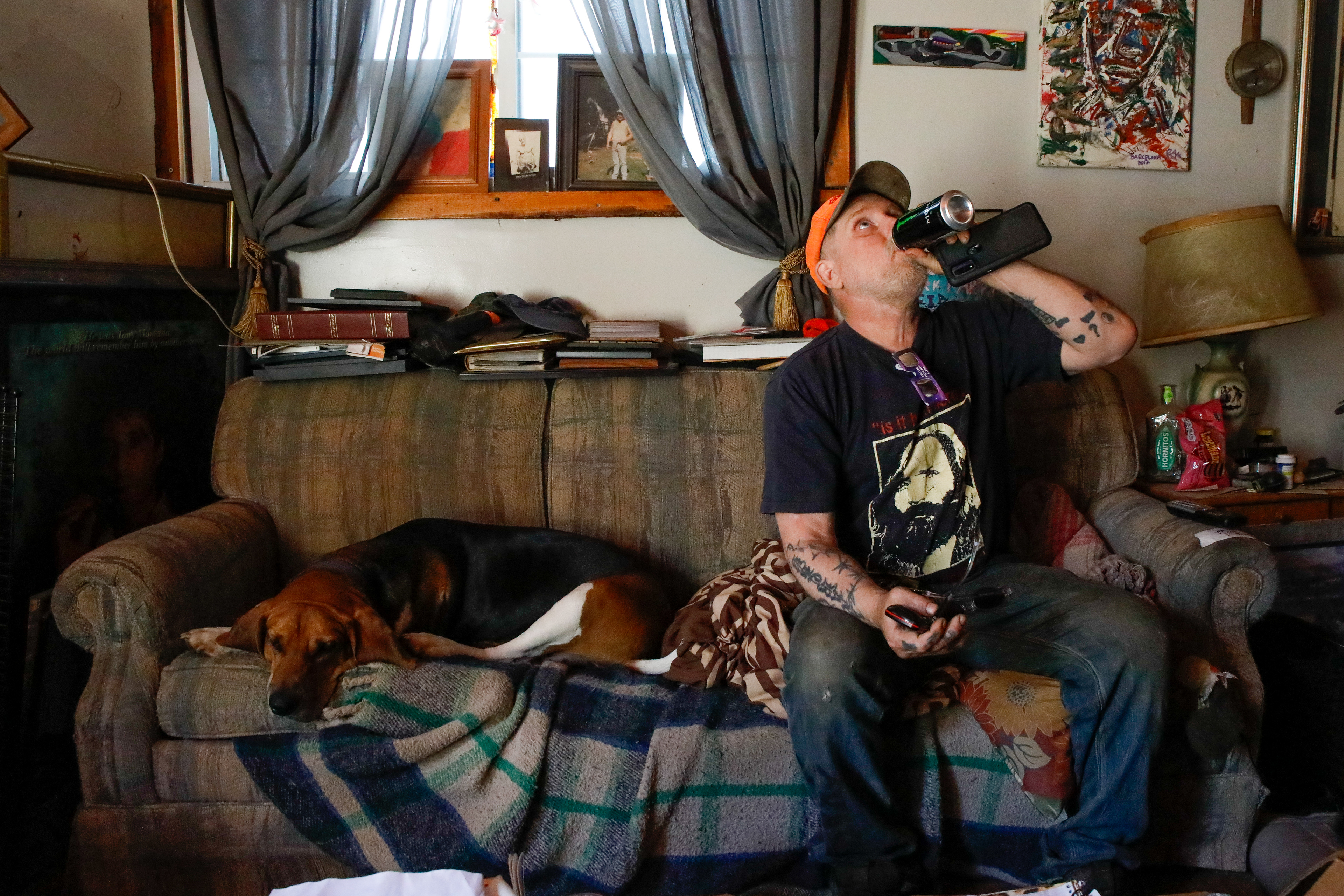 Brewce Martin drinks a Monster Energy drink while sitting on the couch with his dog, Chief, waiting for the North American Solar Eclipse to occur later that day, April 8, 2024. Brewce says he spends a lot of time in this room and with these dogs. When asked what he watches on the TV he says, "I don't know. This shit I guess." He giggles when a Beatles-tribute band is shown singing, "Here Comes the Sun" in reference to the solar eclipse. 