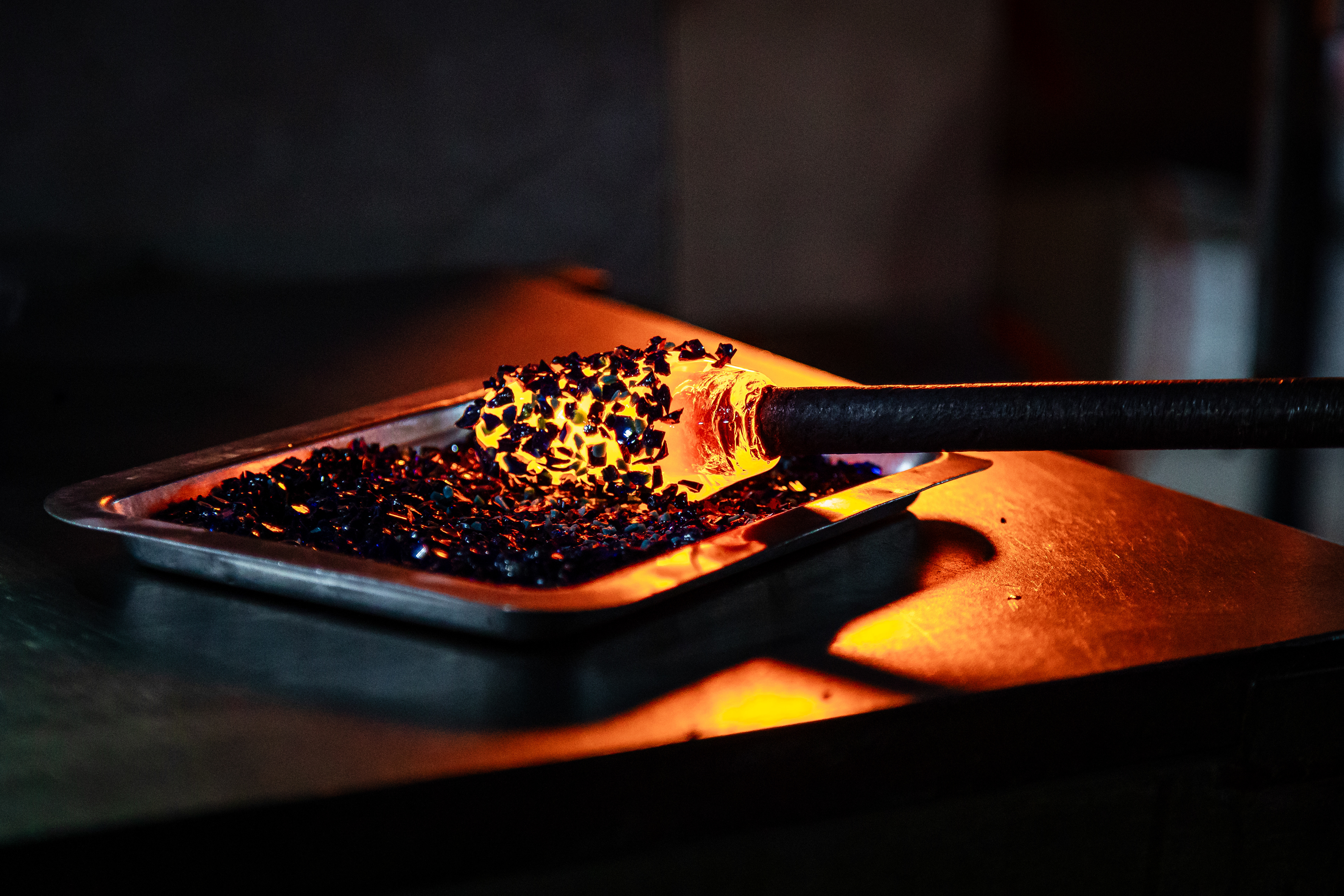 Glass-blowing artists at the Ryukyu Glass Village factory immerse their blowpipes, bearing molten glass, into small colored rock fragments. These fragments give the distinctive vibrant hues to the glass products. The artisans repeat this process until the desired coloration is attained. The craftsmen then will shape the glass by maneuvering the blowpipe or gently blowing into it. These globs of molten glass will later transform into meticulously crafted vibrant glass products.