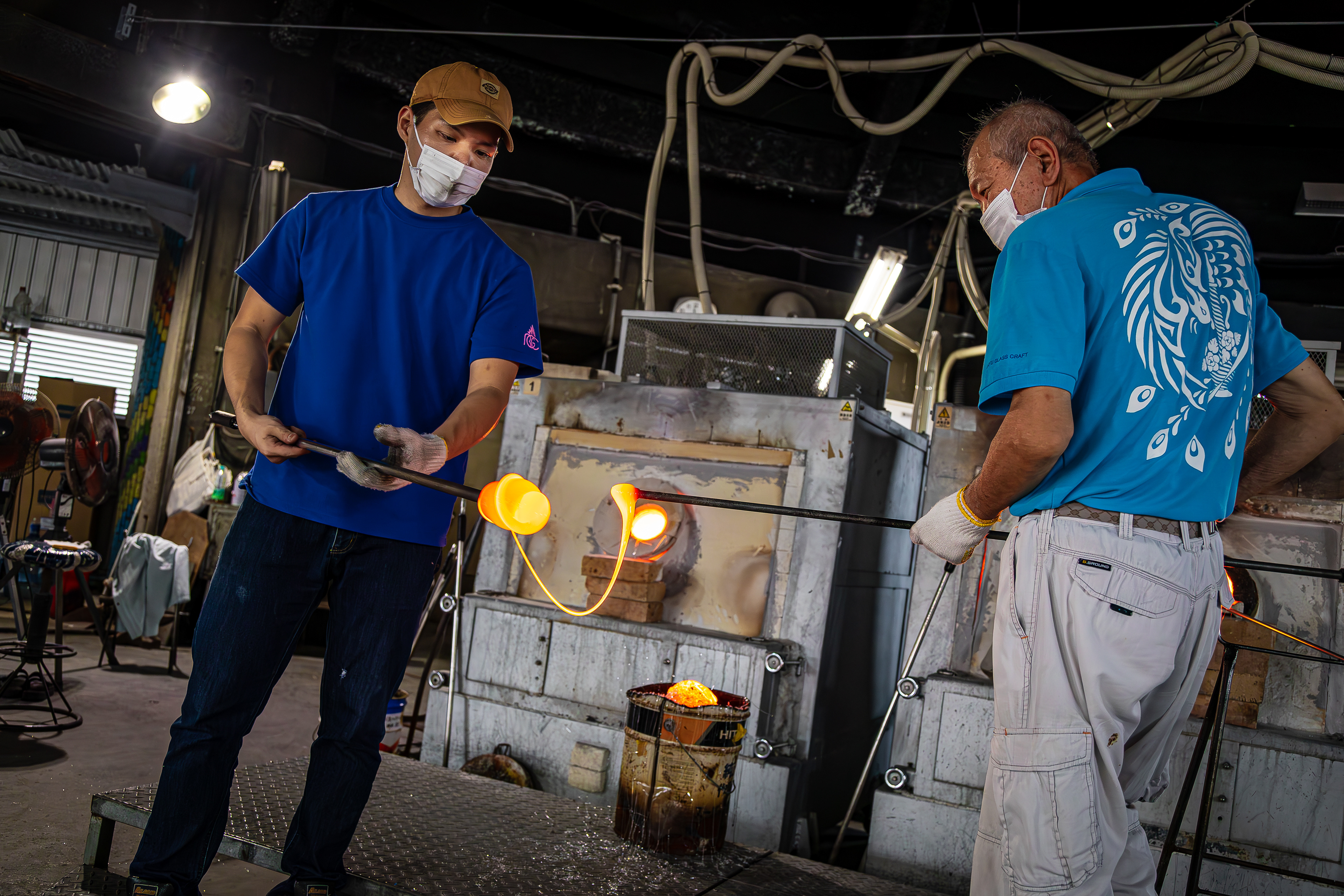 Glassblowing artisans at Ryukyu Glass Village craft vibrant glass products in a workshop, utilizing a kiln reaching 2372 F (1300 C). Amid their creative process, the workshop's internal temperature can soar up to 105 F (40 C). This artistic endeavor involves shaping molten glass by blowing air through a blowpipe, resulting in items such as cups, plates, bowls and more, enhanced with color and meticulous shaping.