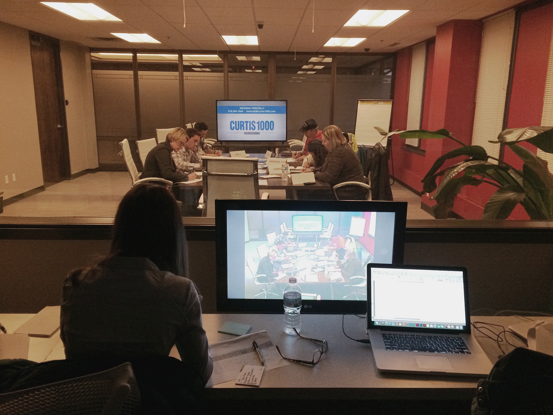 Photograph of a focus group taking place from behind one-way glass. Six participants sit at a table completing a worksheet, a television screen shows the Curtis 1000 logo, and a woman looks on with paperwork and a laptop beside her.