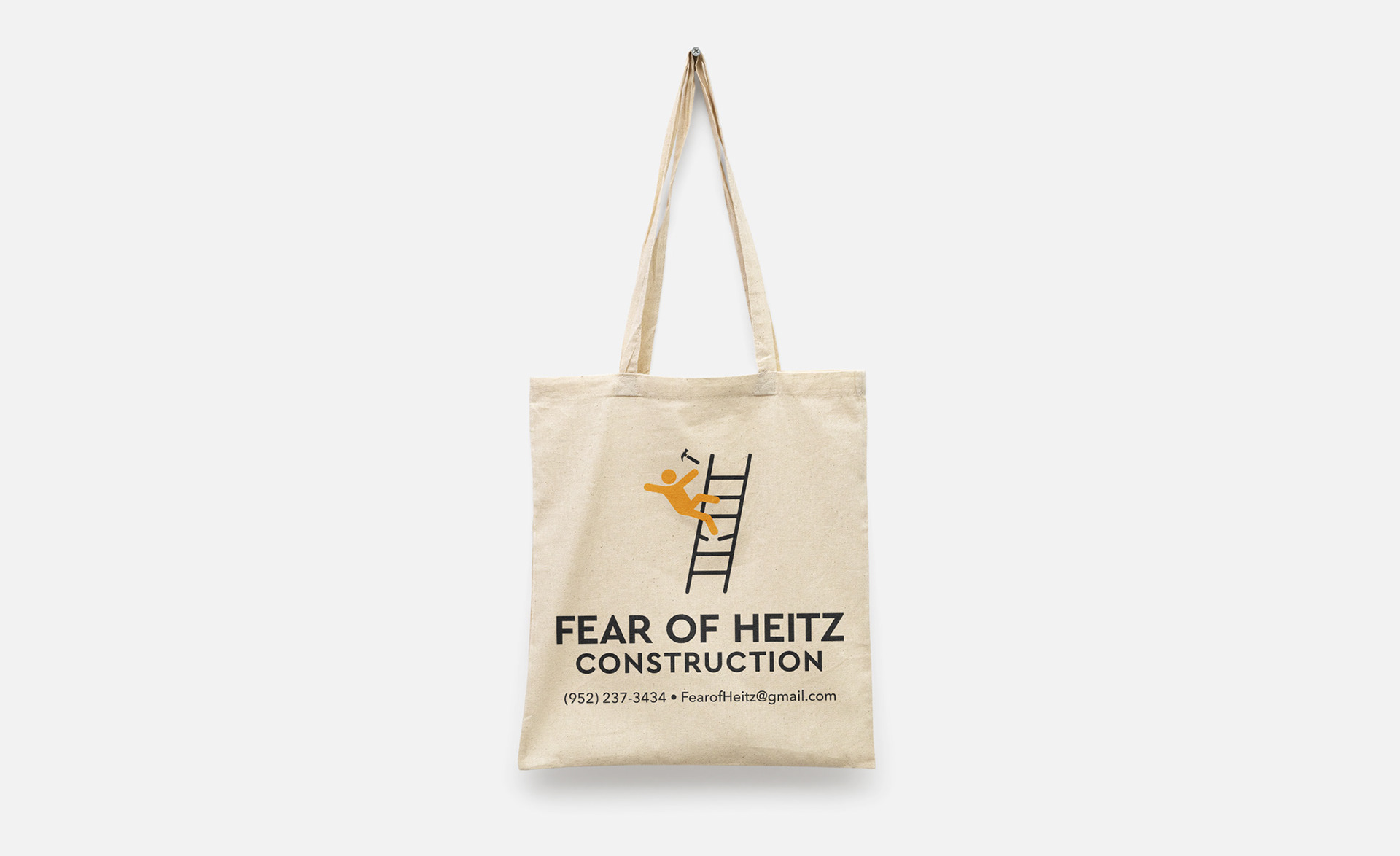 Tan tote bag hung up on a wall with a nail and the text Fear of Heitz Construction with a graphic of a stick figure falling off of a ladder