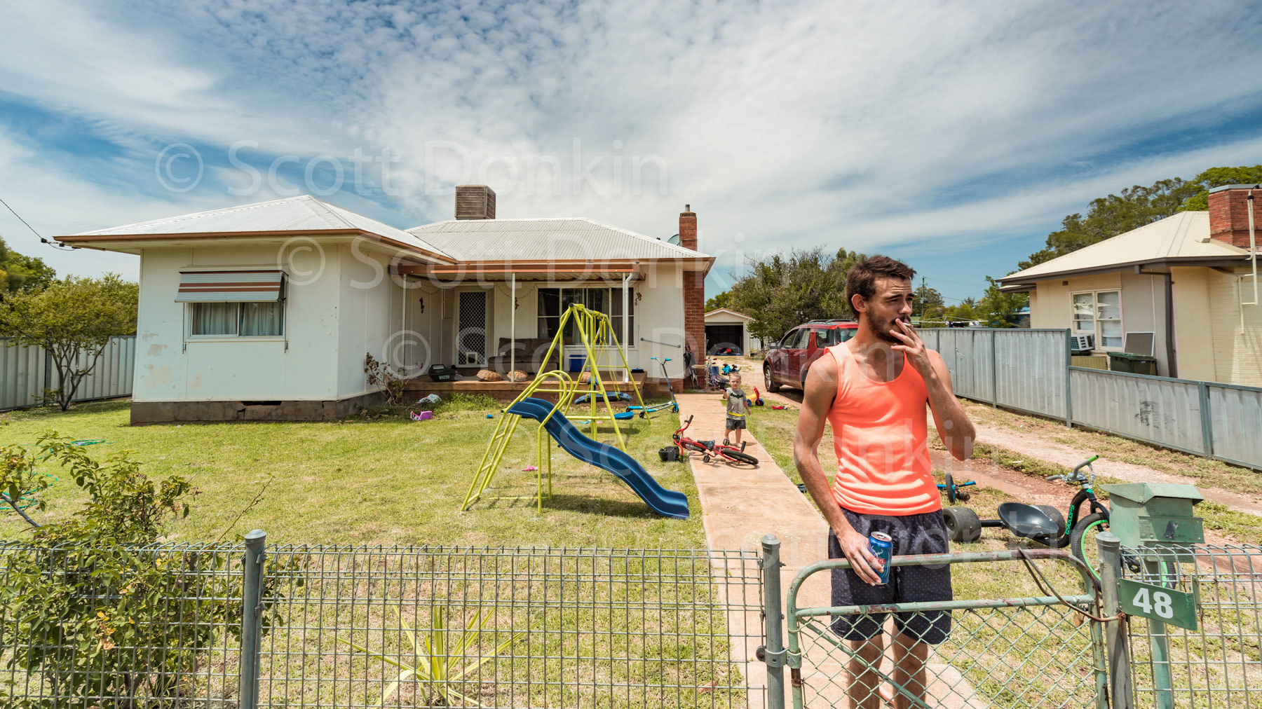 GILGANDRA, NSW, AUSTRALIA - 1 February 2019: A man stands at his front gate having a drink and a cigarette after work.  The town of Gilgandra is located 460 Km northwest of Sydney and services the surrounding agricultural area where wheat is grown extensively together with other cereal crops.  The surrounding farms are also known for their sheep and beef cattle.