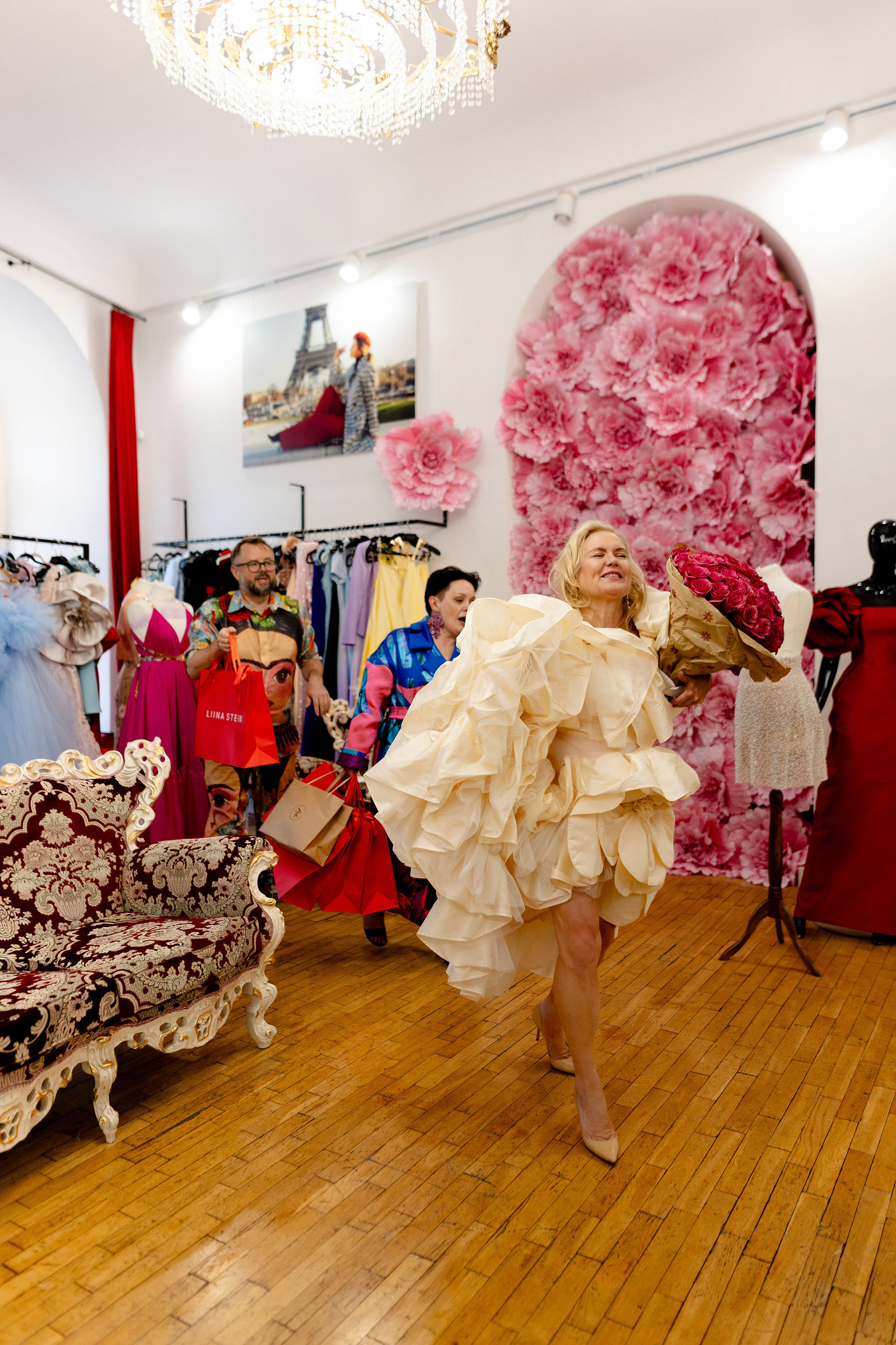 Osaleja valges Liina Steini couture-kleidis lahkub salongist lillekimbuga, taustal värvikad kleidid ja salongi interjöör.