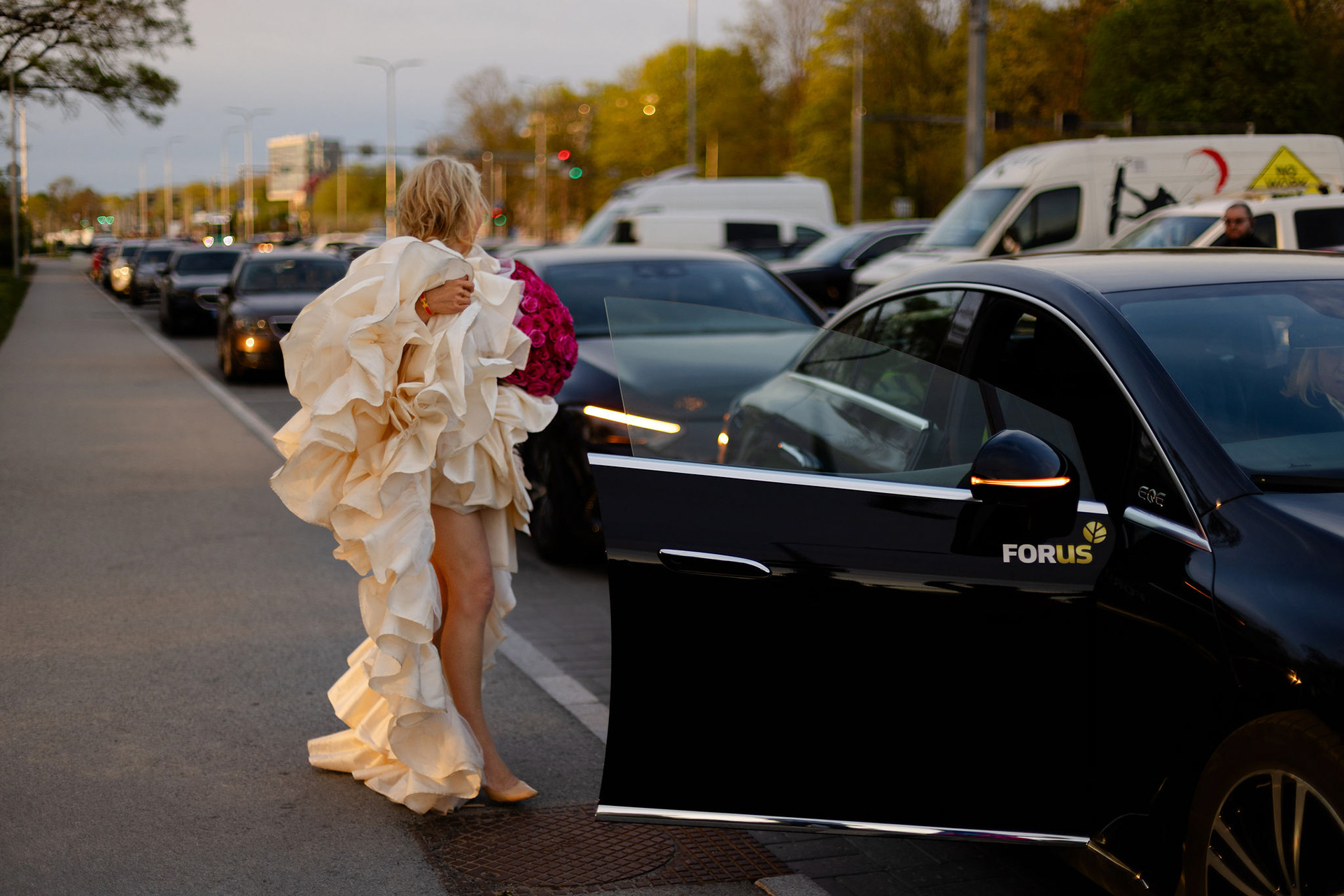 Naine valges Liina Steini couture-kleidis ja roosa lillekimbuga siseneb õhtul autosse pärast inspireerivat "Uued algused" fotosessiooni Tallinnas.