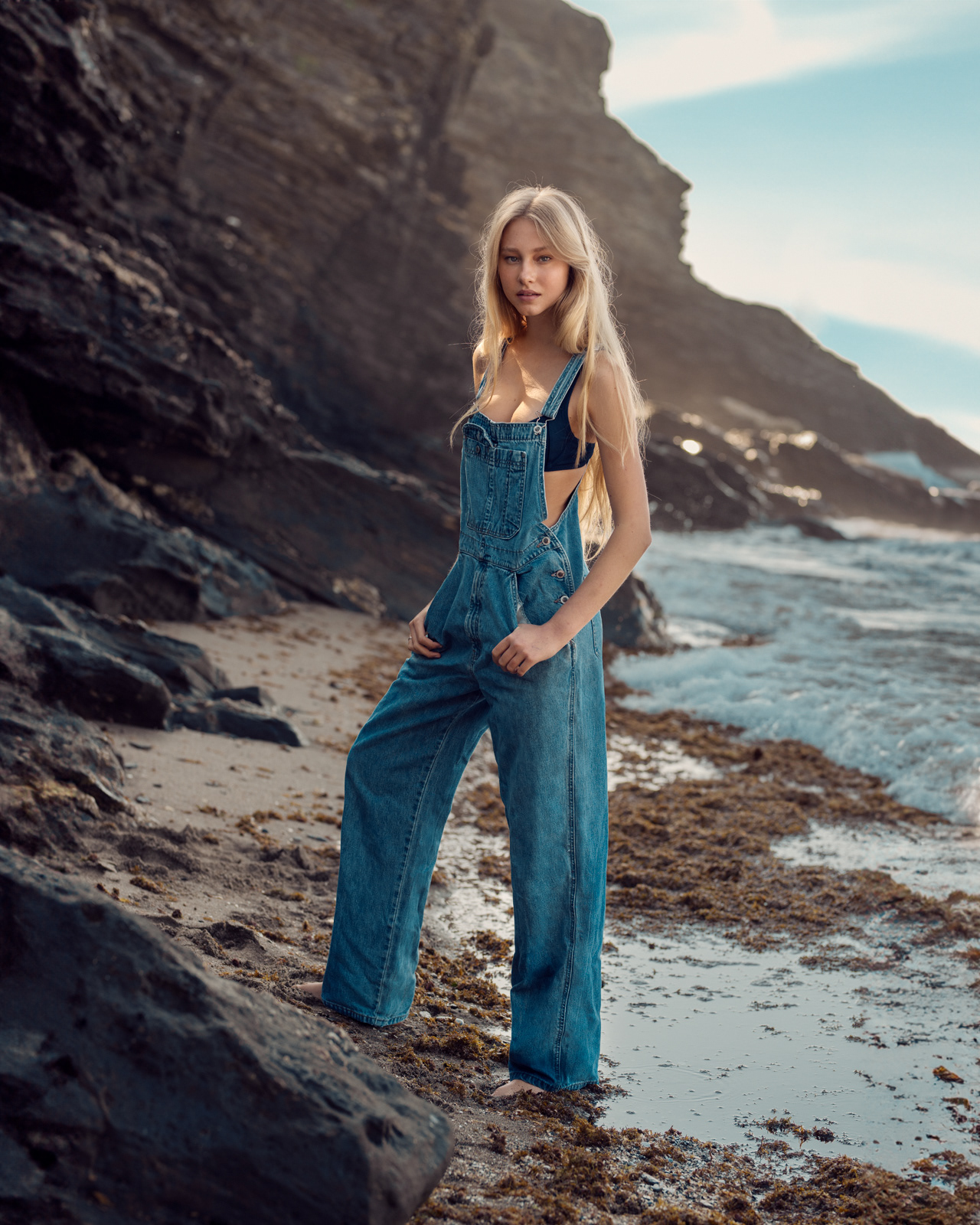Outdoor portrait of model in denim overalls standing on rocky shoreline – Joanna Kustra lifestyle fashion photography