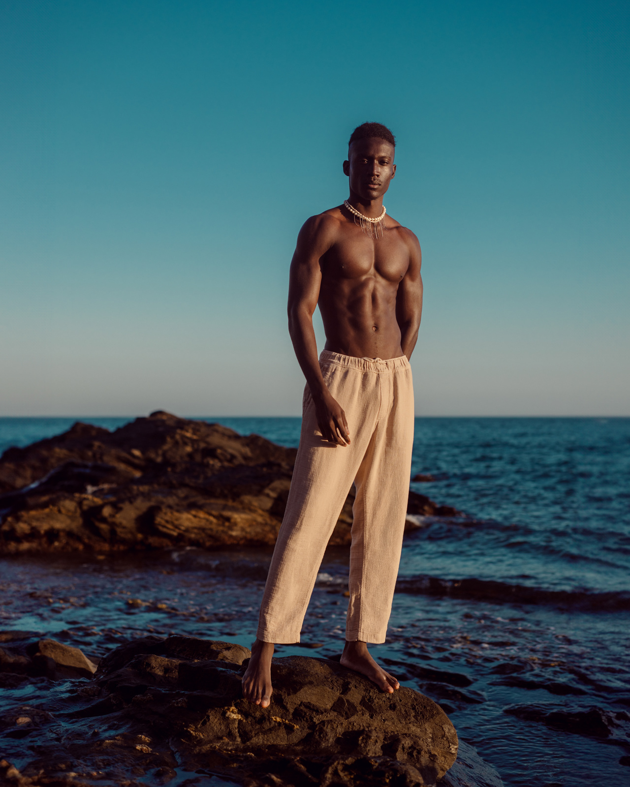Male model standing on rocky shoreline in beige trousers – Joanna Kustra men’s fashion photography
