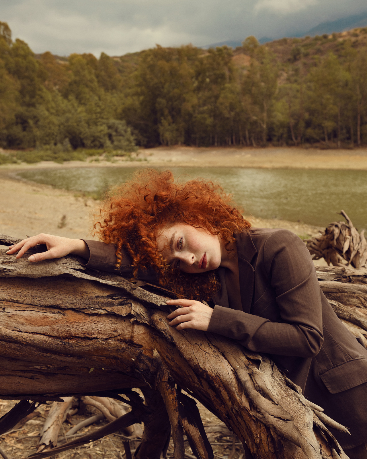 Outdoor fashion portrait of red-haired model leaning on driftwood by the shore – Joanna Kustra, natural light photography