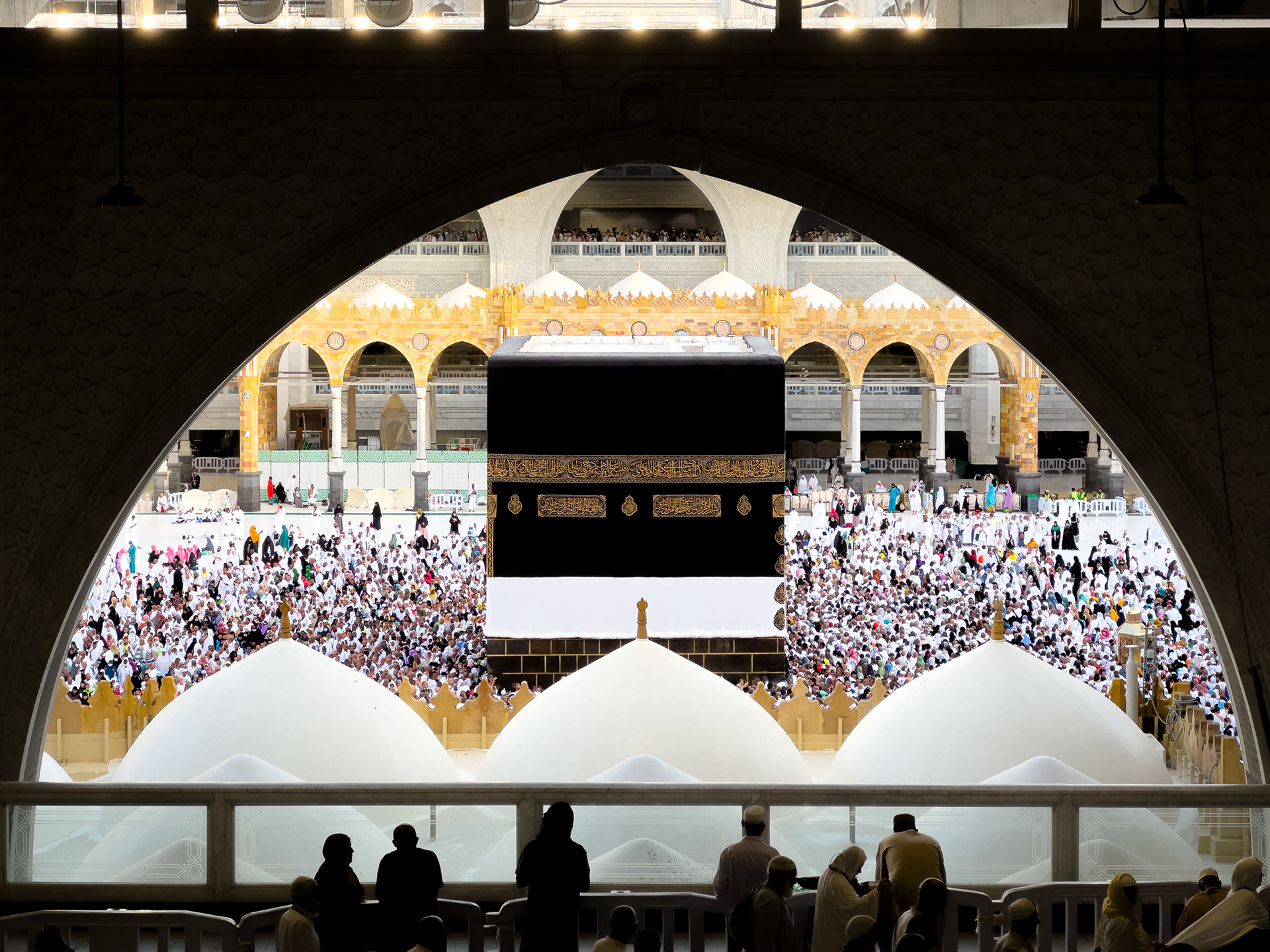 Masjidil Haram, Mecca, Saudi Arabia — The Kaabah fills the archway, draped in the Kiswah, surrounded by thousands in white. Silhouettes in the foreground watch in stillness. The scale of the scene — the vastness of the courtyard, the smallness of each individual — only becomes apparent when you stop and look properly.