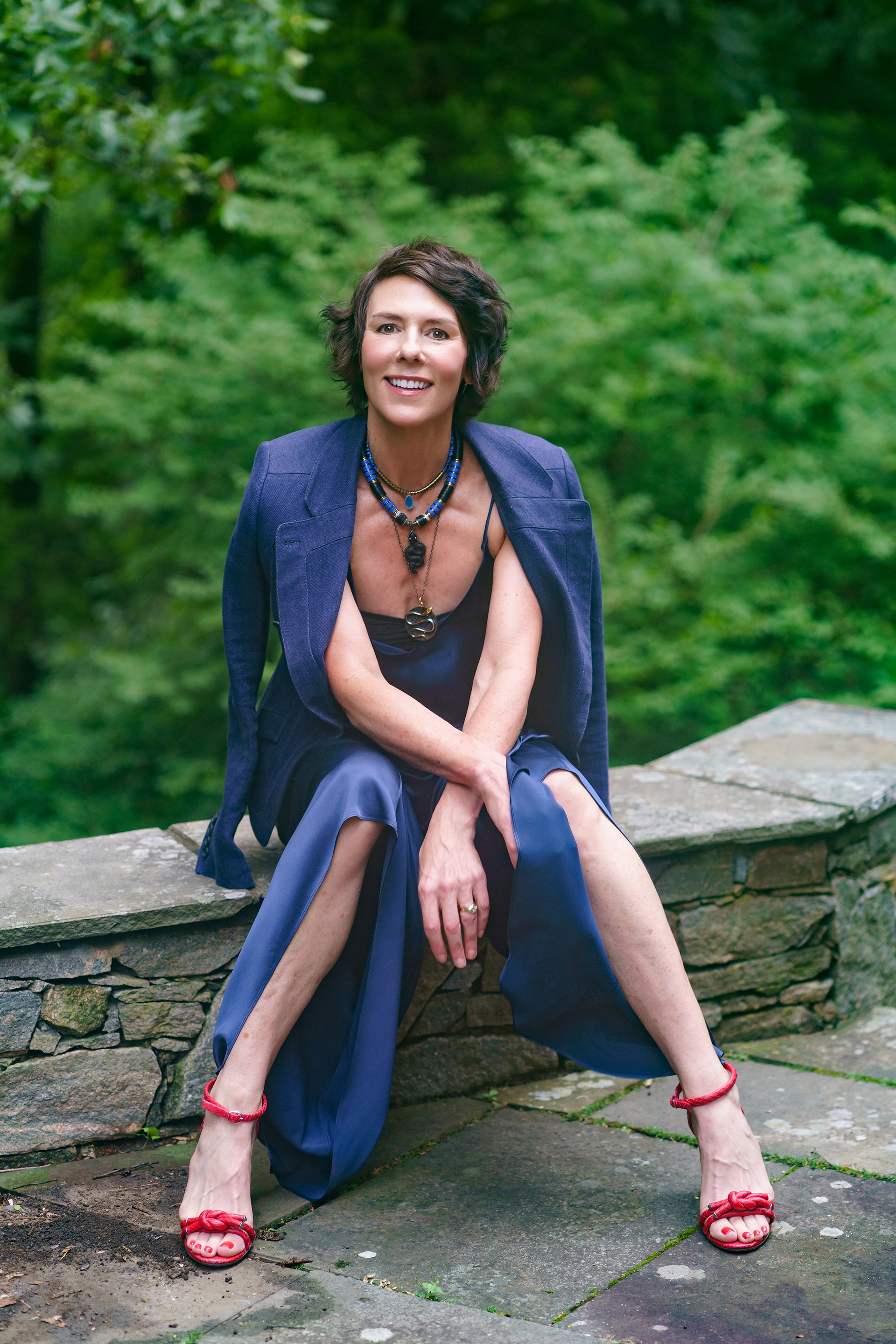 Sarah Easley of Maison Marche wearing a navy blue silk dress with blazer covering her shoulders, posed sitting on a rock ledge wall with greenery in background