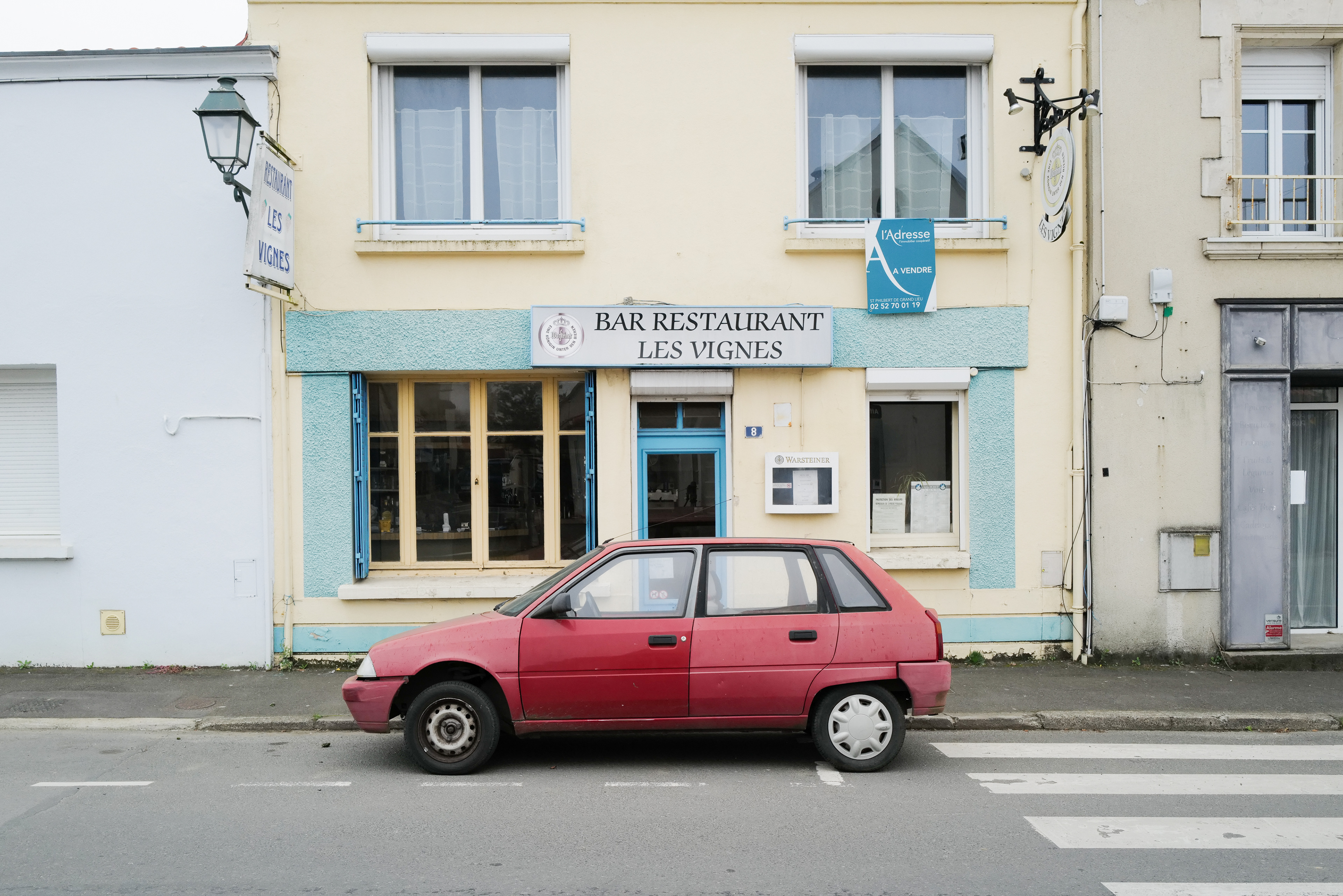 Une vieille AX rouge aux couleurs lessivées est garée devant un bar restaurant appelé "les vignes" et donc le bâtiment est à vendre.