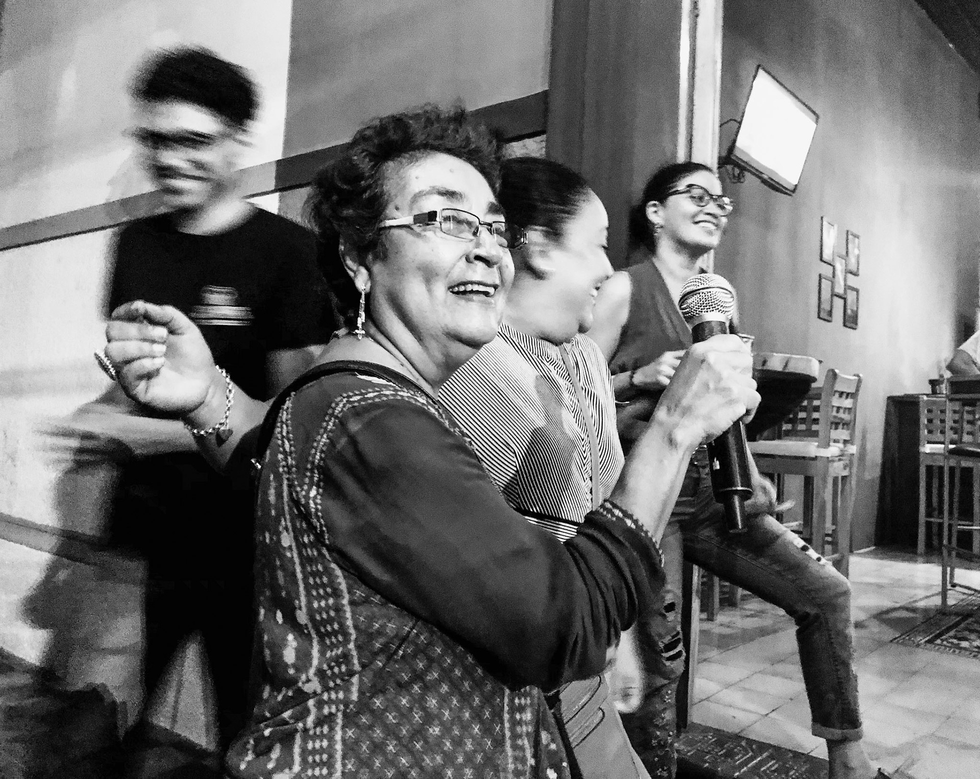 An older woman sings into a microphone with a joyful smile during a lively karaoke night, surrounded by friends who laugh and cheer her on.