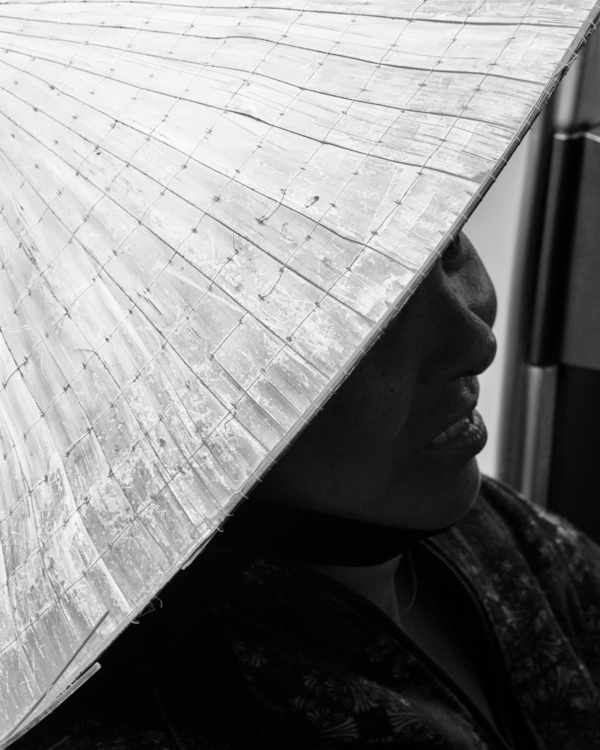 A woman under a nón lá, Biên Hòa — caught while walking the streets. The hat's shape goes back over 2,500 years; bronze figures wearing them show up in ancient rain ceremonies for the harvest. Palm leaf stitched over bamboo rings, tilted just enough to leave half a face in shadow.