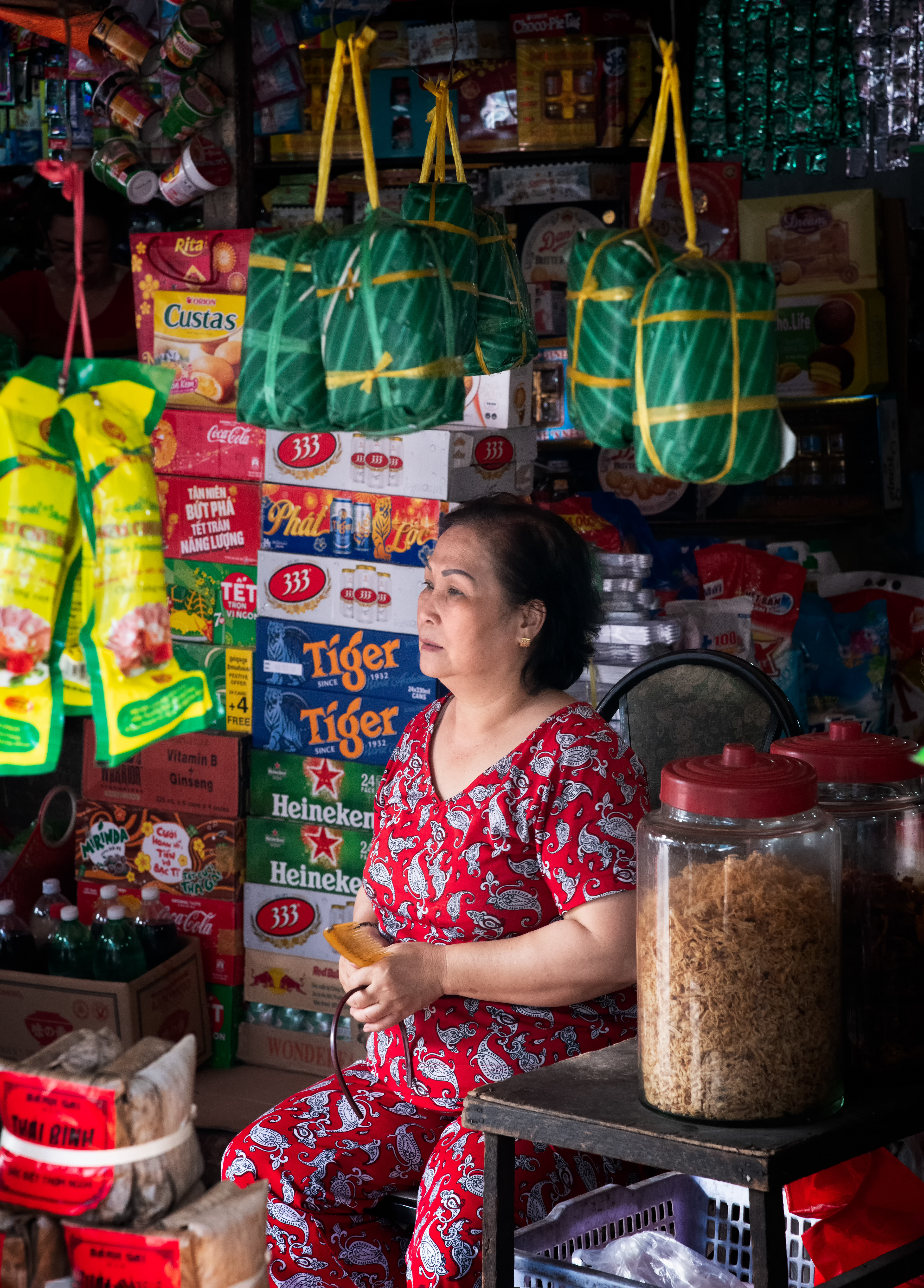 A shopkeeper on the road, Biên Hòa — bánh tét hanging behind her, sticky rice and pork wrapped in banana leaf, boiled half a day. Best known as Tết food, but eaten here year-round. In the south they say the cylinder shape stands for the sky, the way the north's square version stands for the earth.